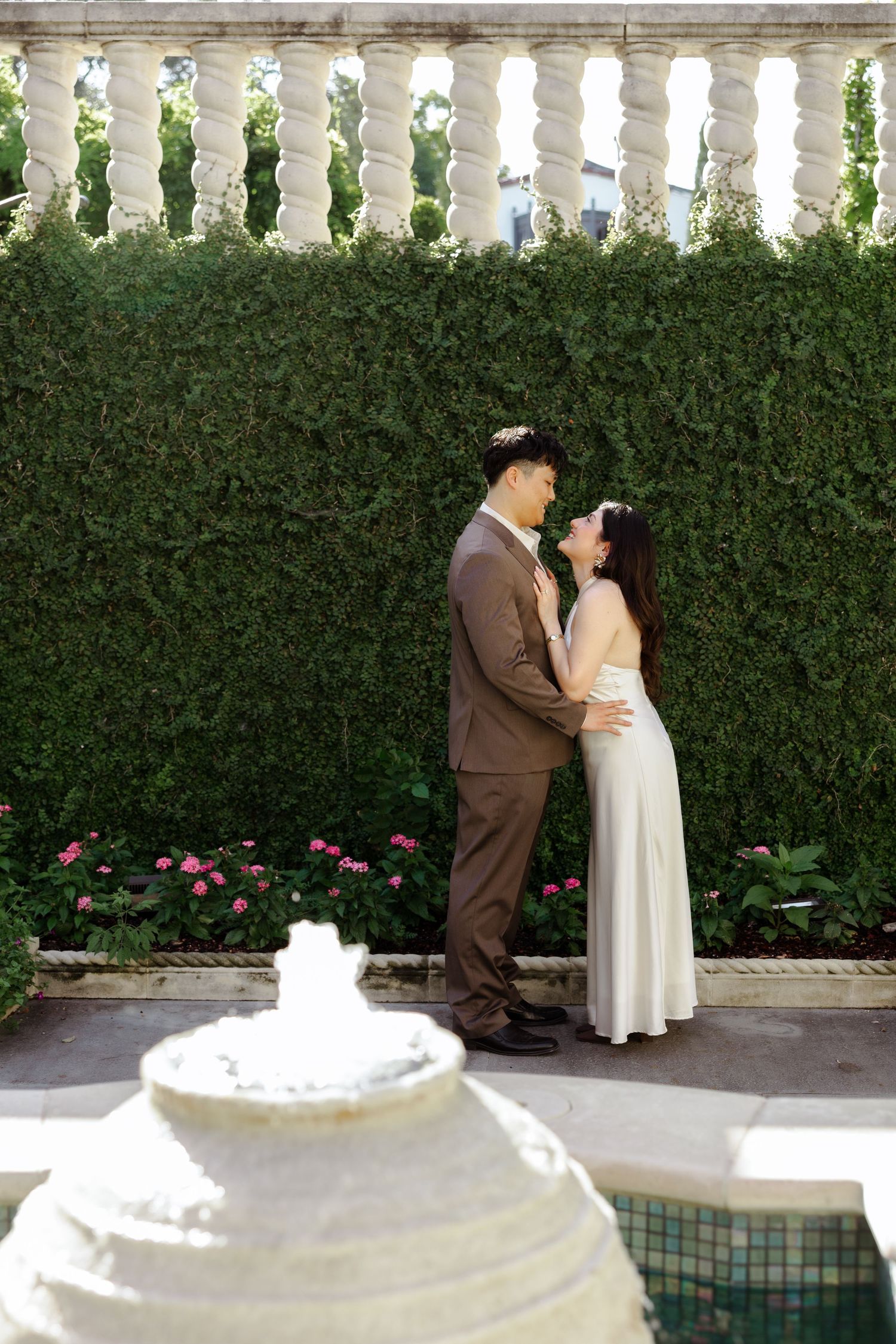Couple in wedding attire embracing before a lush green hedge wall with pink flowers and a stone balustrade.