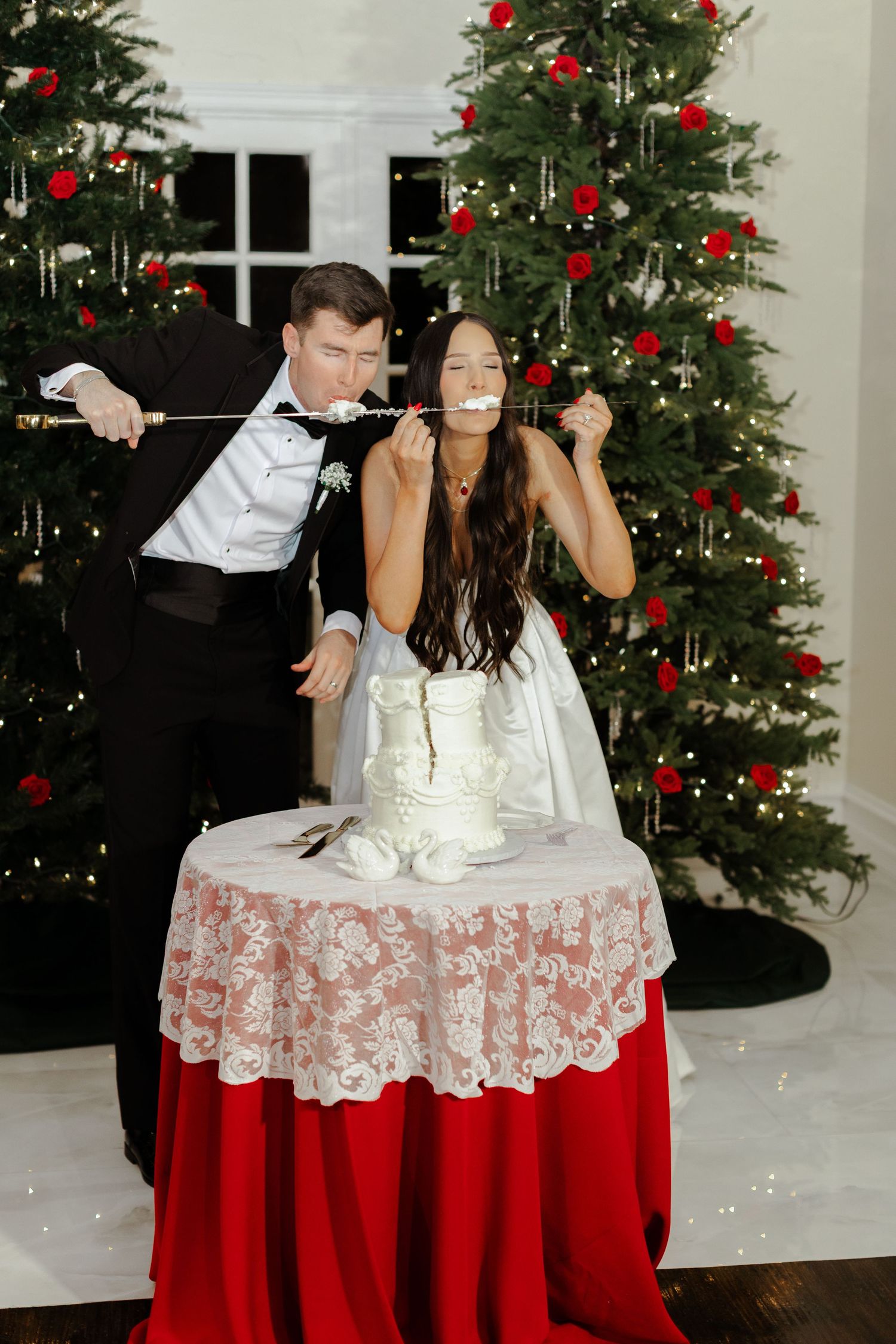 A couple poses playfully on a red and white tablecloth in front of a decorated Christmas tree with red ornaments.