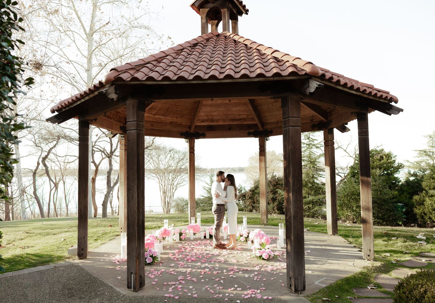 A couple stands under a wooden gazebo in a park with pink cherry blossom petals scattered on the ground.