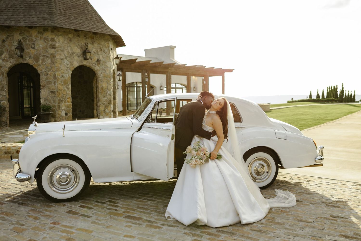 Bride and groom kissing beside a classic white vintage car at an elegant outdoor wedding venue.