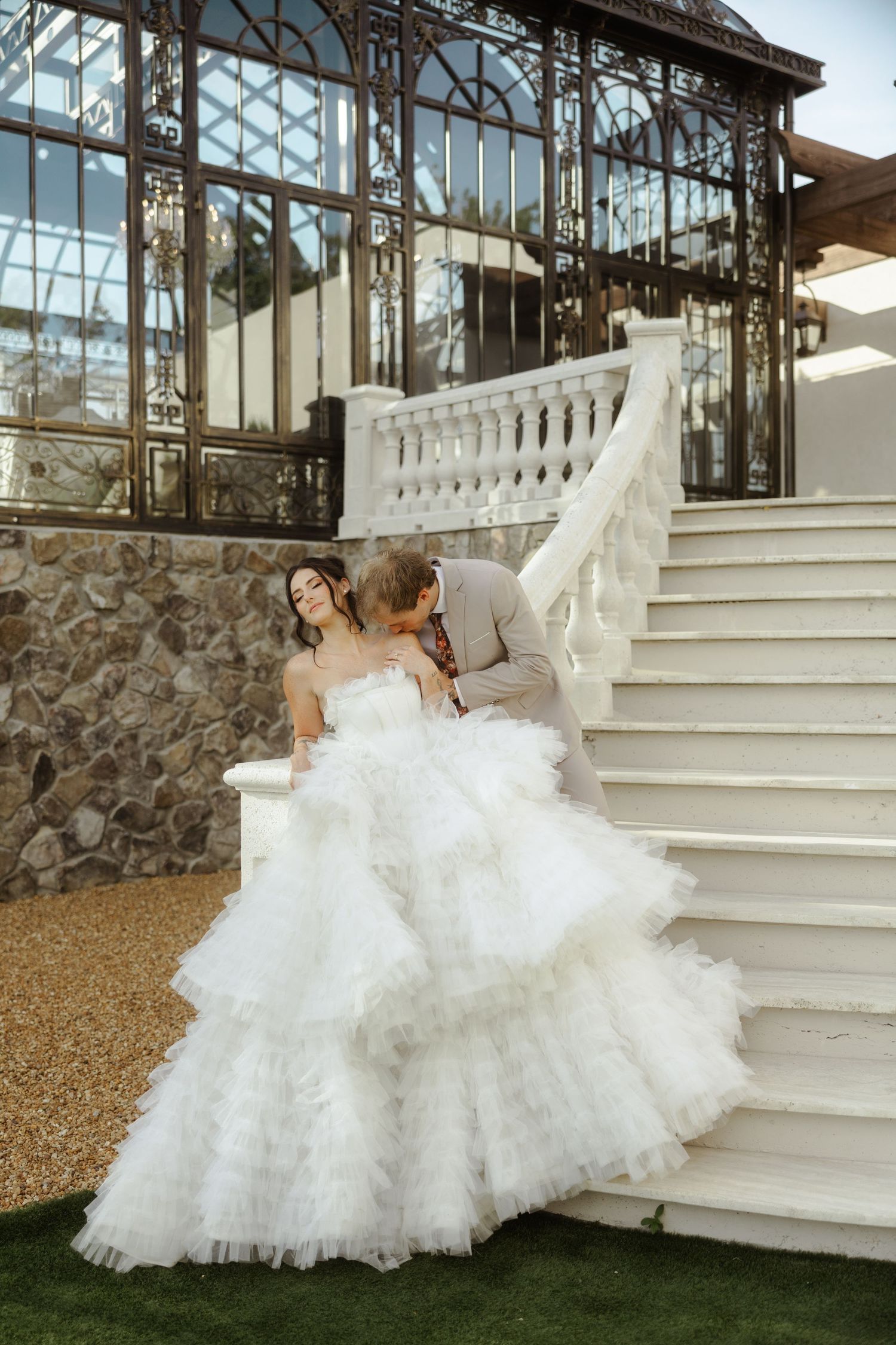 Bride in layered white tulle gown with groom in gray suit sharing a romantic moment on white stairs.