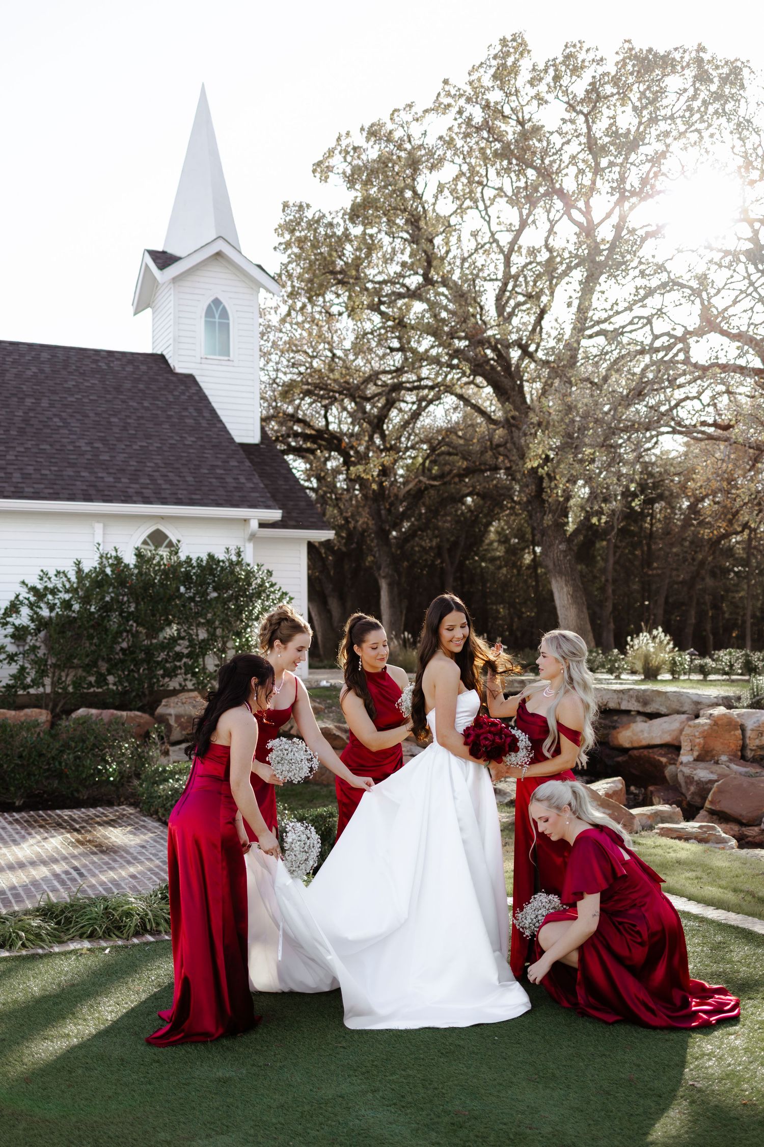 Bridesmaids in red dresses help arrange a bride's gown outdoors near a white chapel.