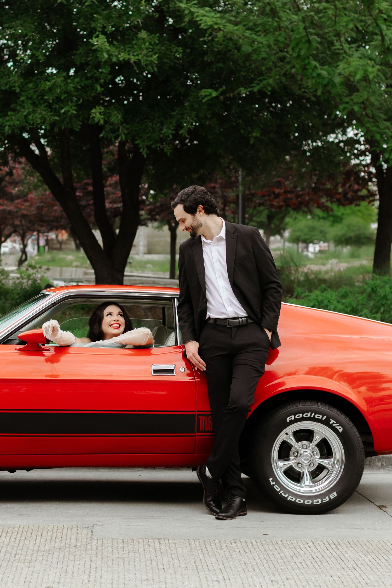 A woman in a red classic muscle car smiles at a man in a black suit leaning against the car outdoors.