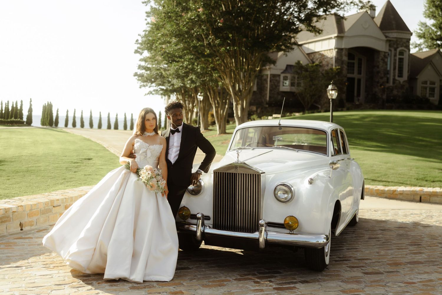 Bride and groom posing beside a classic white Rolls-Royce on their wedding day at an elegant estate.