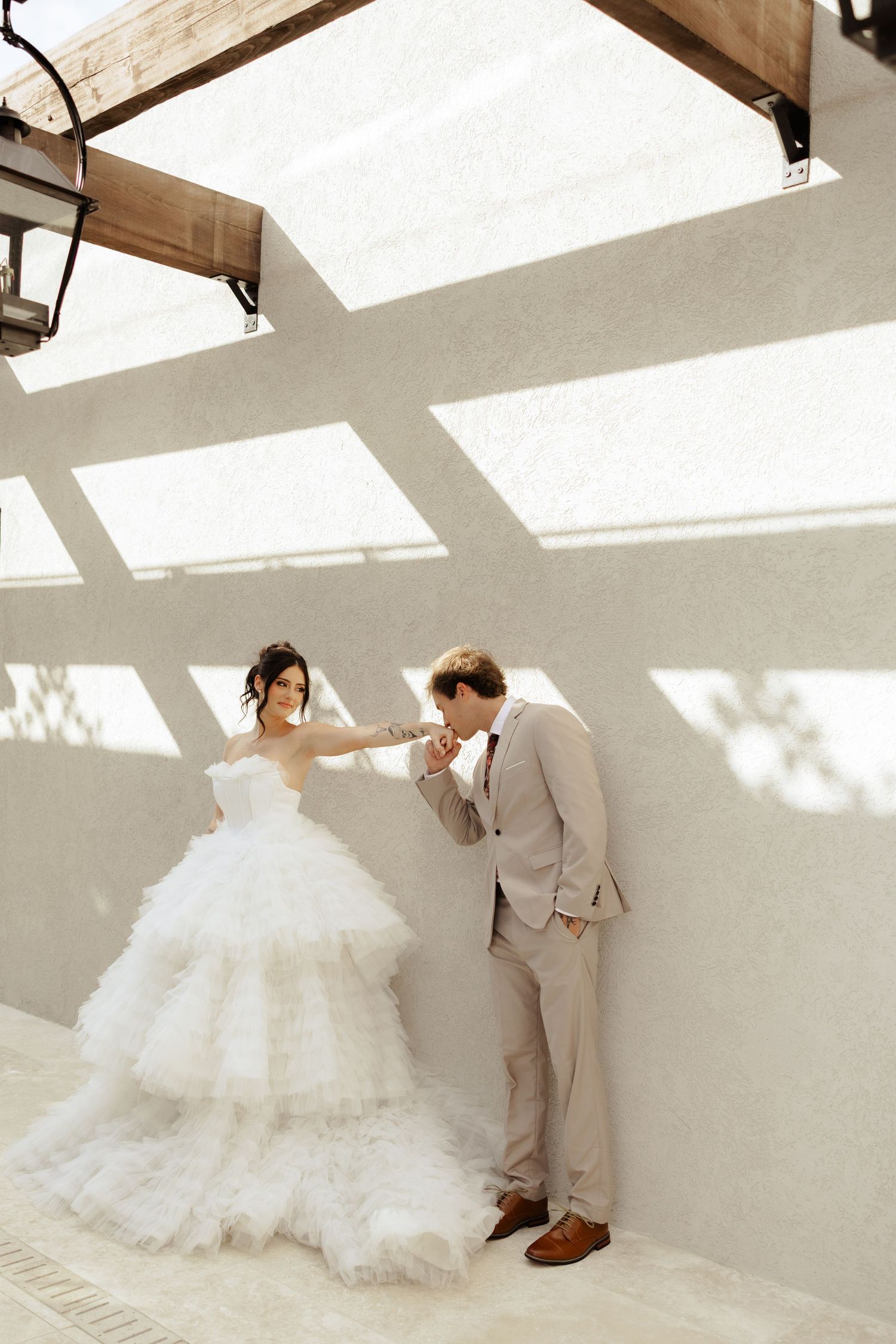 Groom in beige suit kissing bride's hand, bride in layered white tulle ballgown, standing against sunlit white wall.