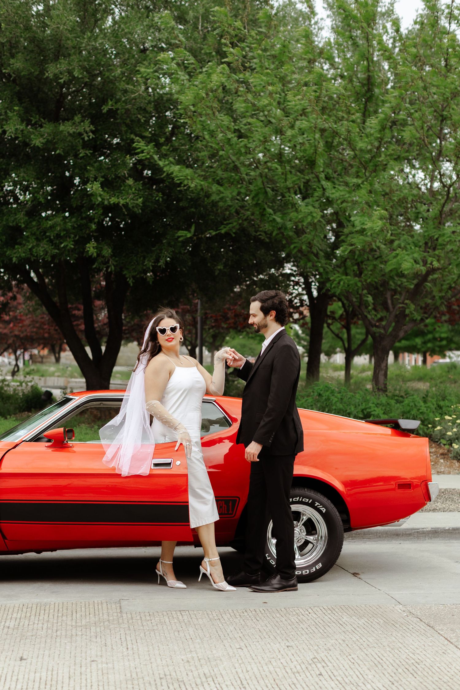 Bride and groom posing beside a classic red muscle car outdoors, bride wearing a white jumpsuit and veil with sunglasses.