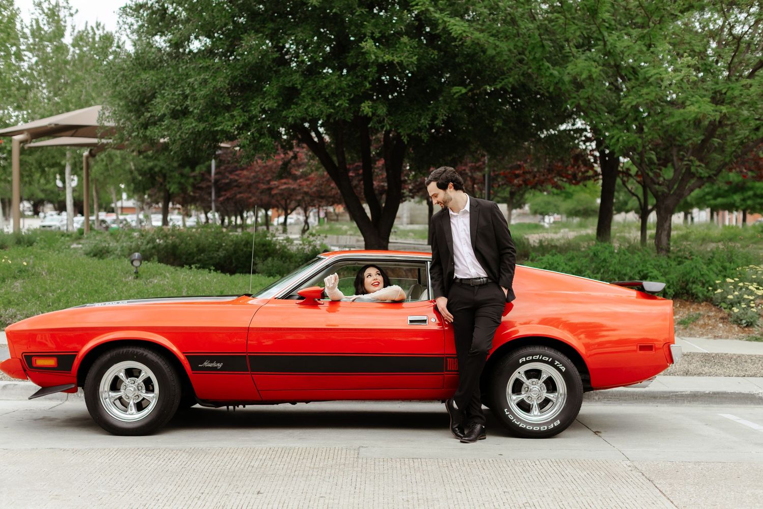 Couple poses with a classic red Ford Mustang, man in black suit leaning on car door while woman sits inside.