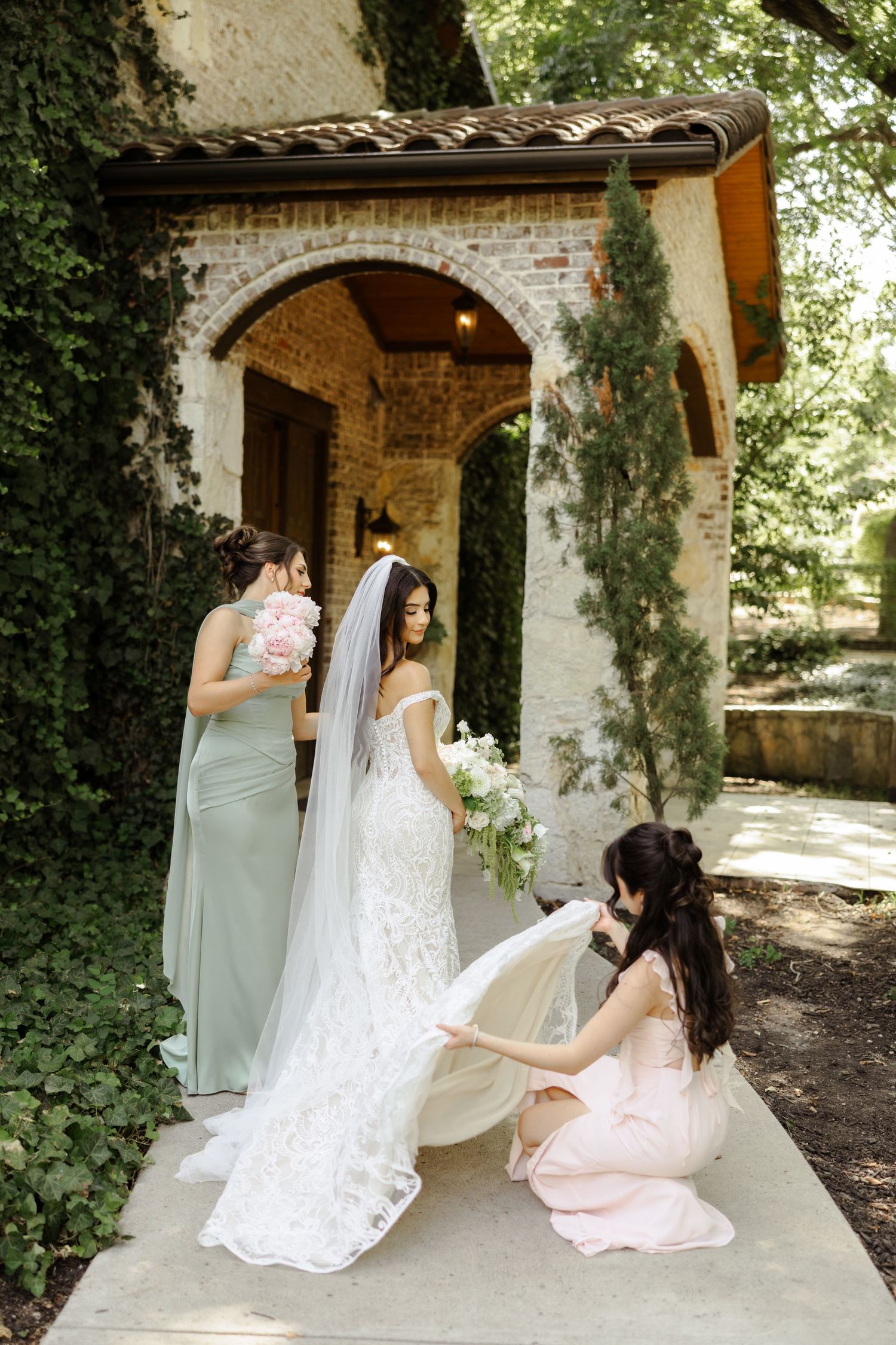 Bridesmaids in sage and blush gowns help bride with lace wedding dress outside rustic brick chapel.