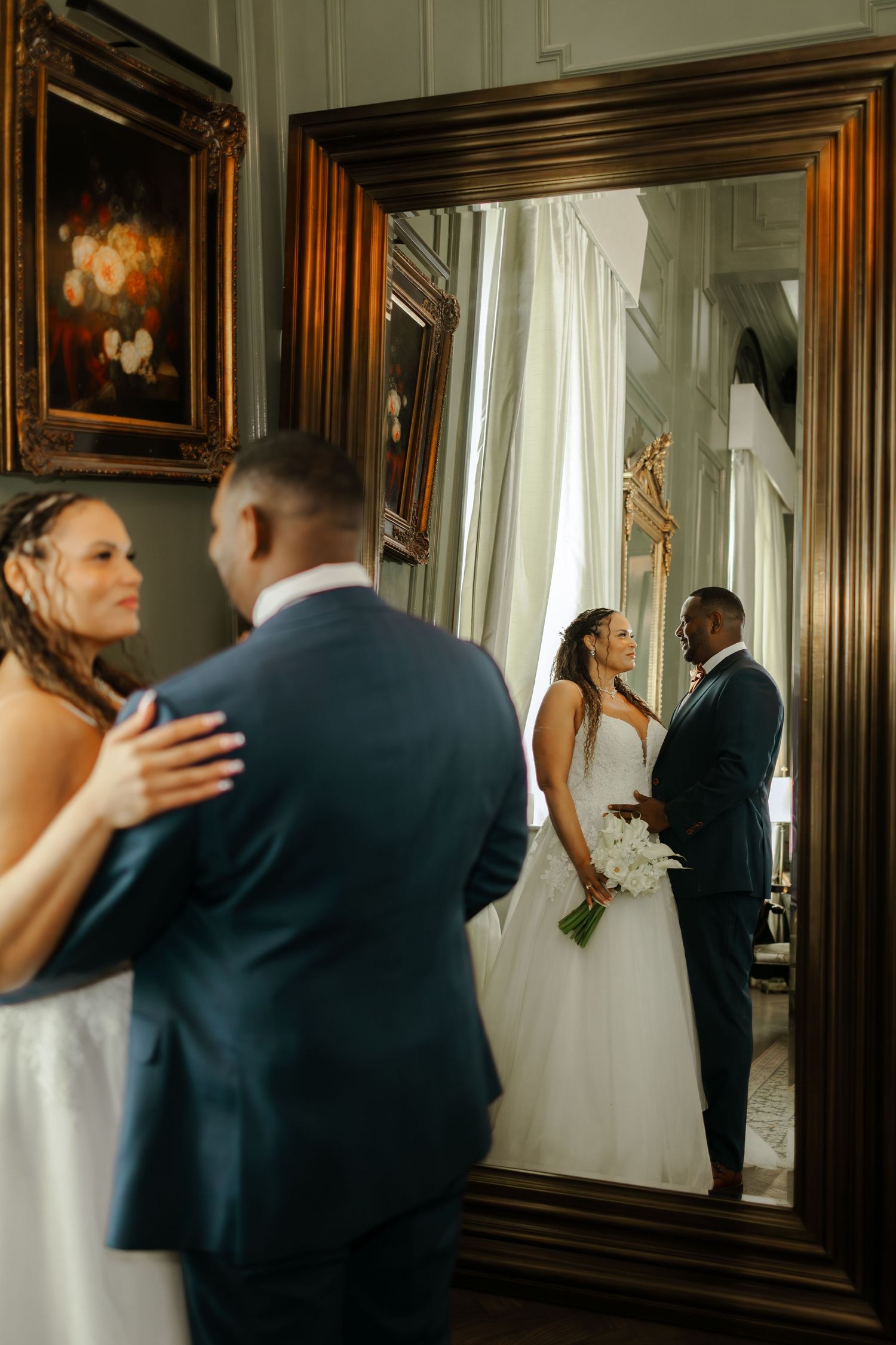 Bride and groom share a romantic moment reflected in an ornate gold mirror at their elegant wedding venue.