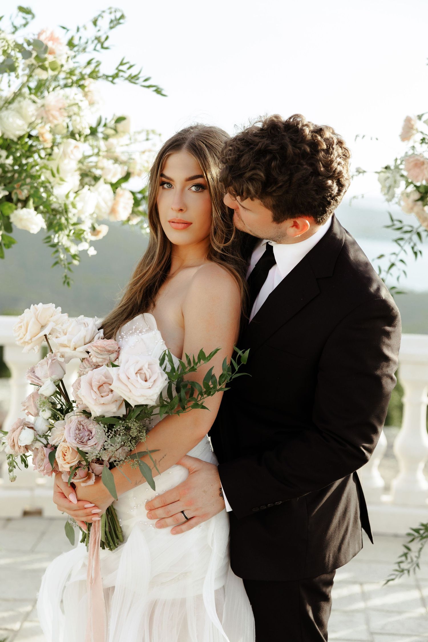 Groom in black suit kisses bride holding blush bouquet on scenic outdoor terrace with mountain views.