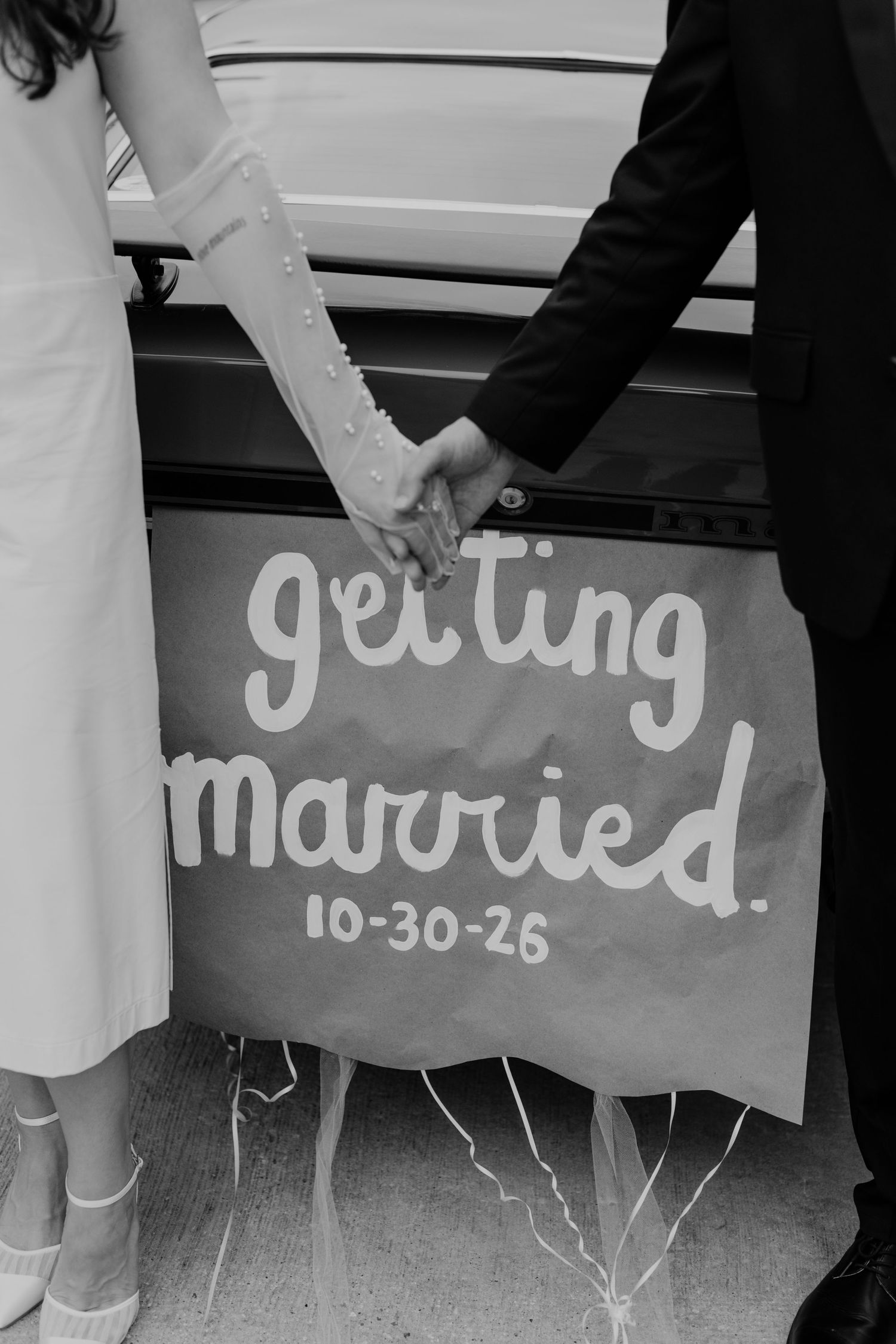 Black and white photo of couple holding hands beside a 'Getting Married 10-30-26' sign on a car.
