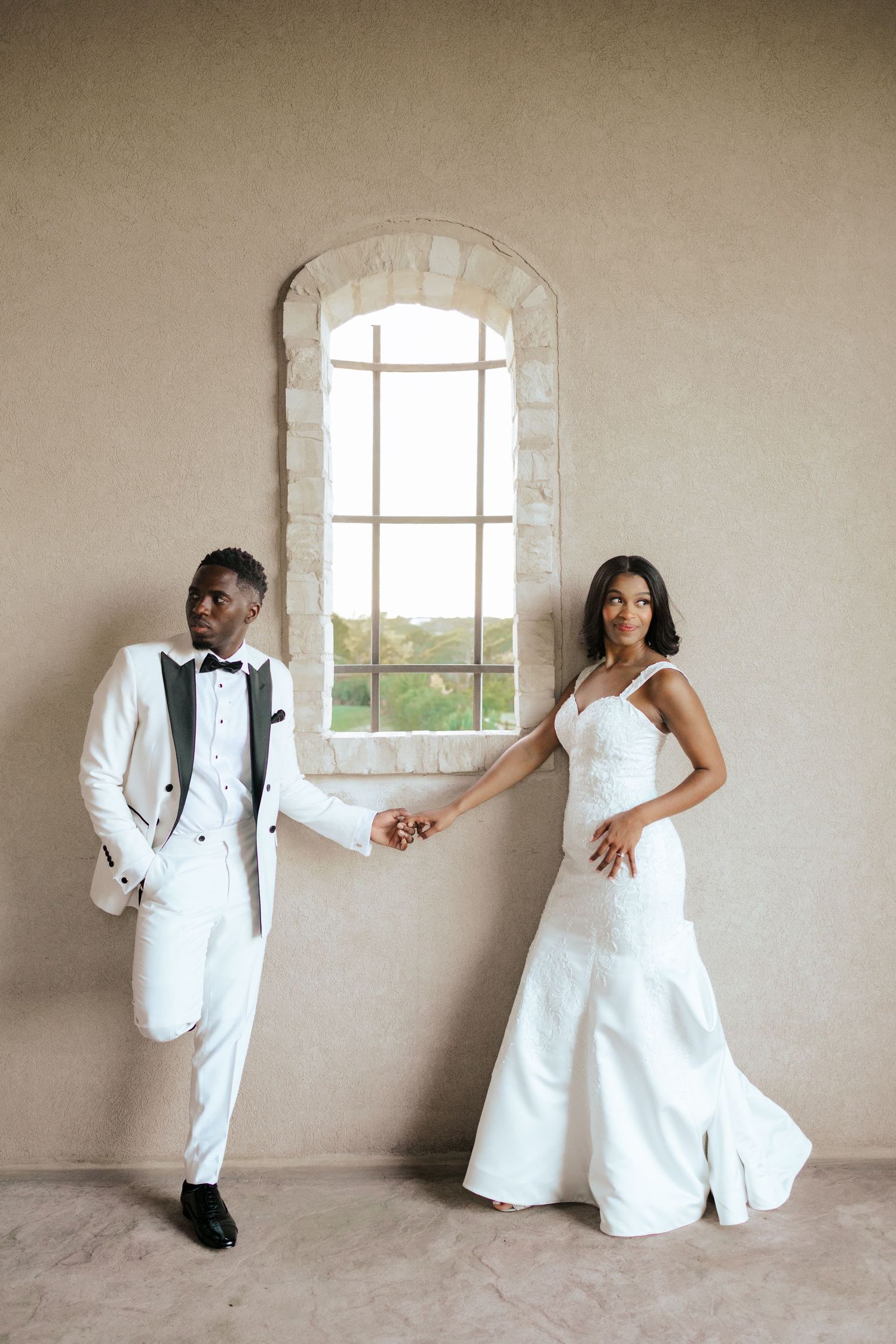 Couple in matching white attire poses playfully against neutral wall with arched window.