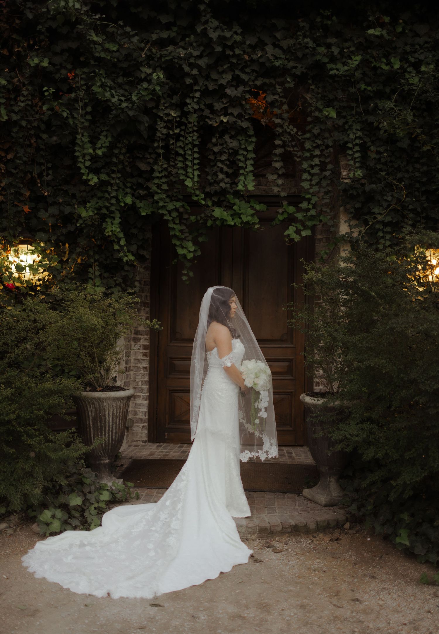 A bride in a flowing white wedding dress and veil stands in front of a rustic brick wall surrounded by greenery.