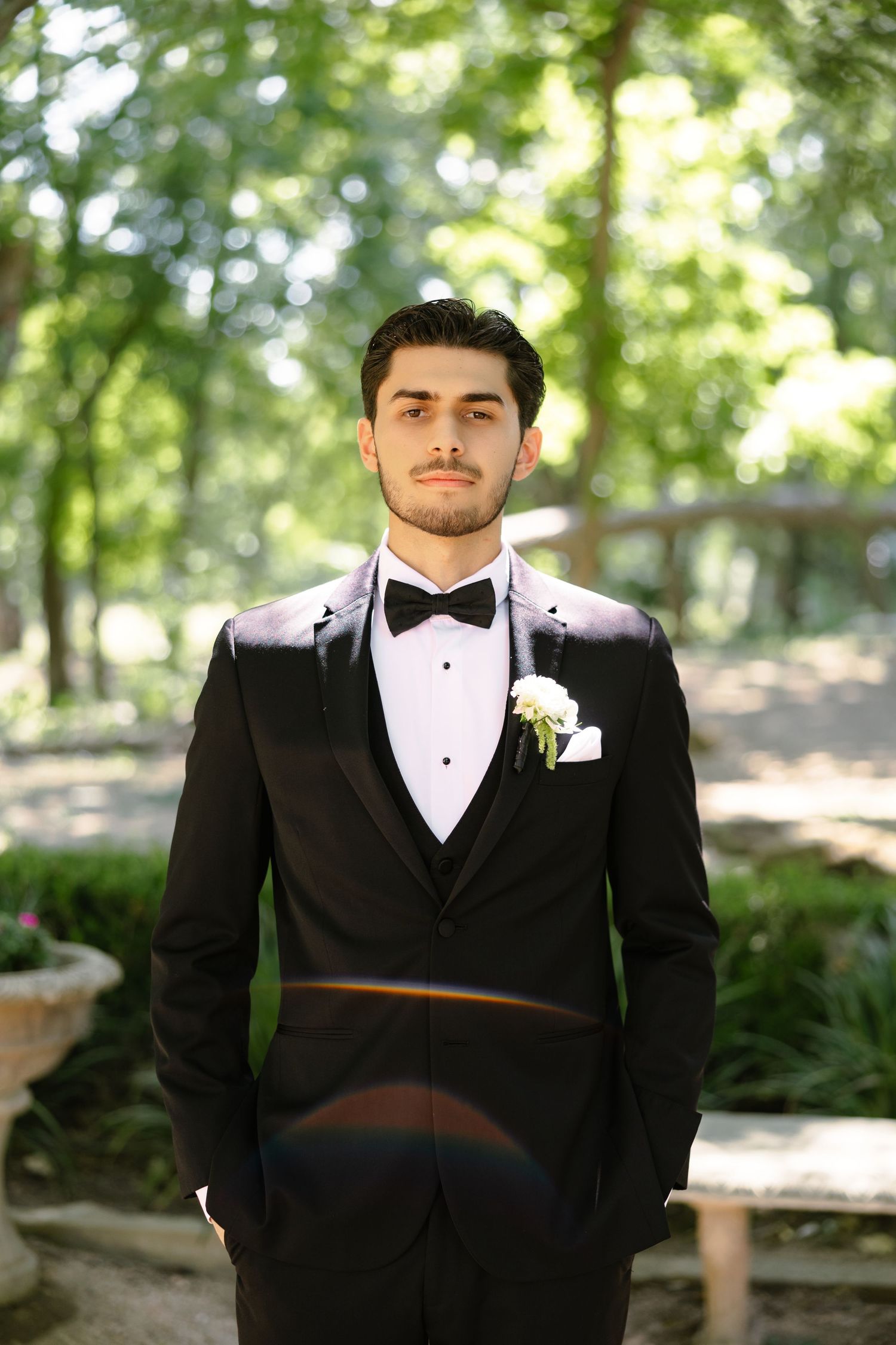 Elegant groom in black tuxedo with bow tie and white boutonniere posing outdoors in a lush green garden setting.