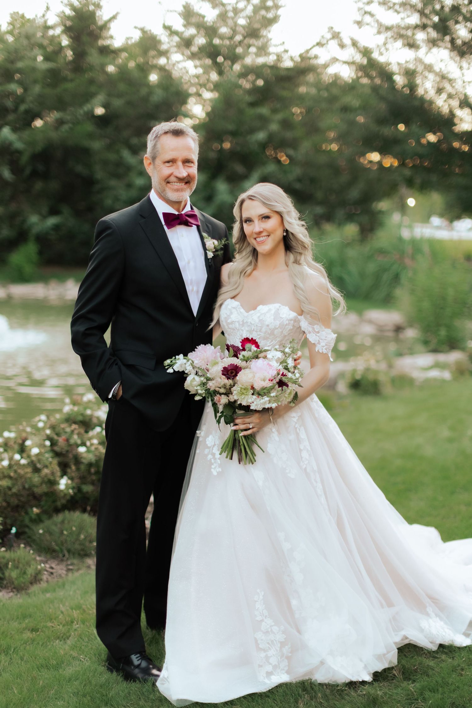 A wedding couple poses together in formal attire with bouquets in a garden setting at sunset.
