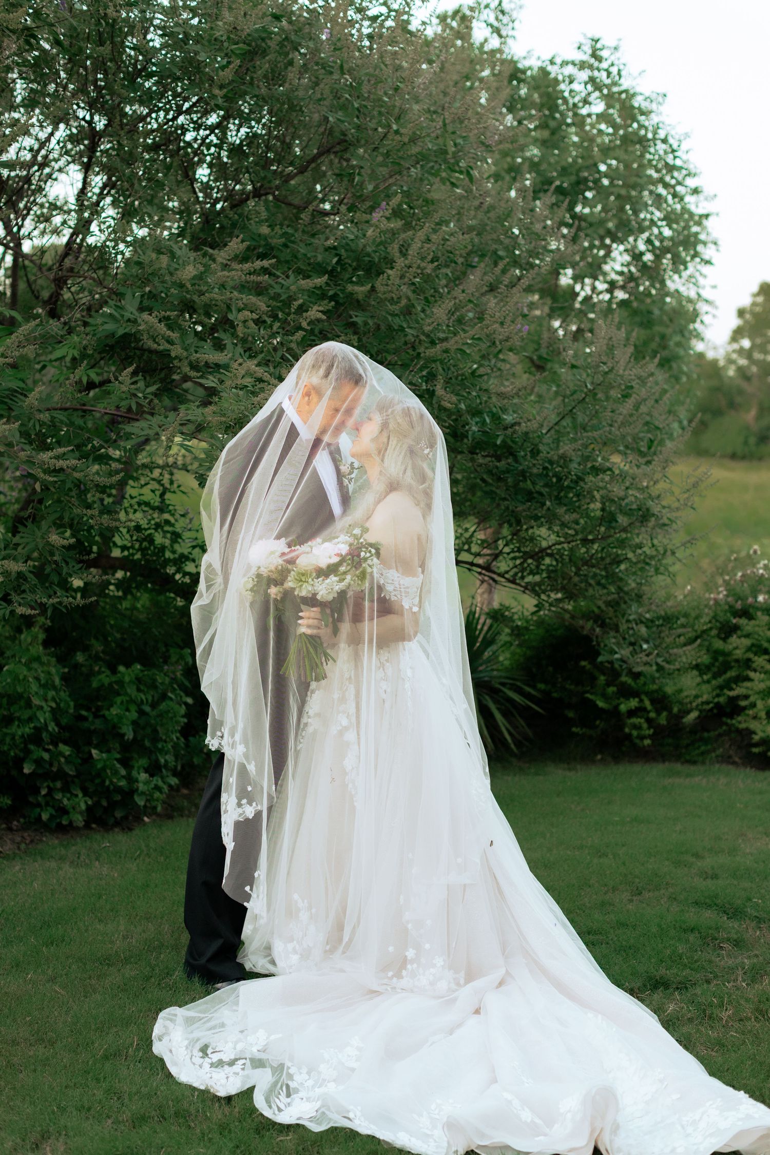A romantic wedding moment captured under a veil with flowing train in a garden setting.
