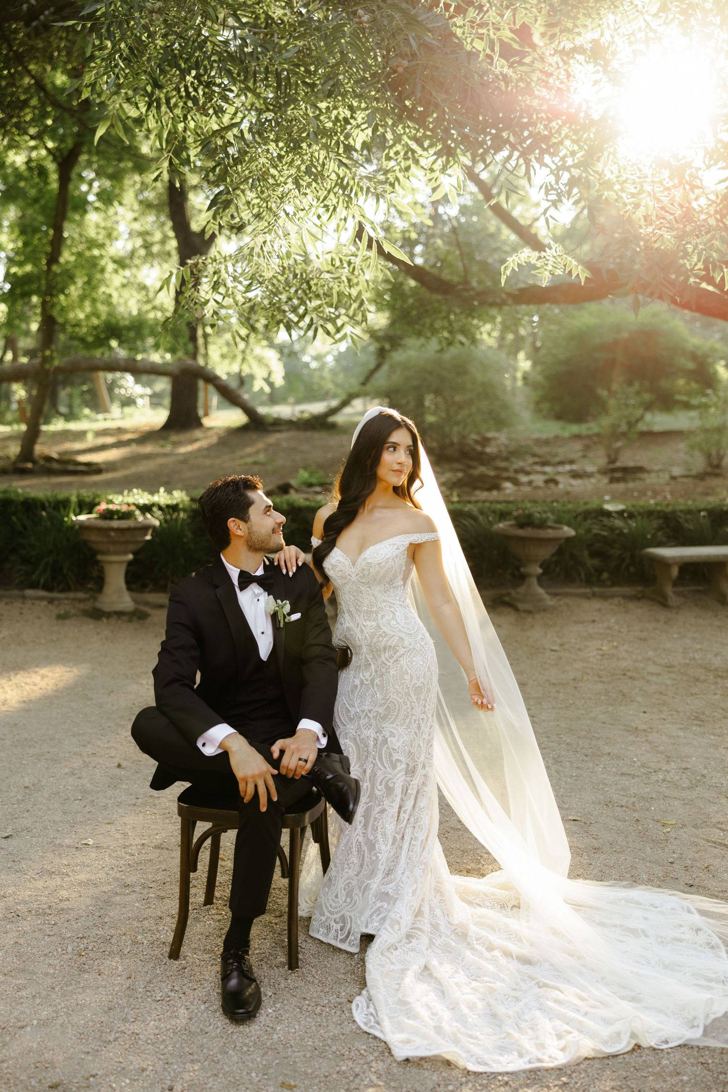 Bride in lace gown with veil beside groom in tuxedo seated outdoors under sunlit trees at garden wedding.