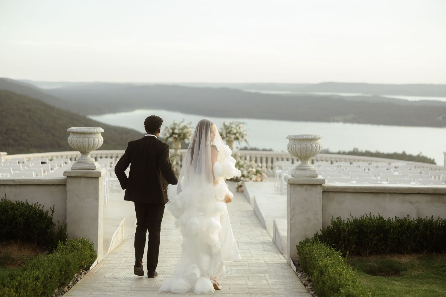 Bride and groom walking hand in hand on elegant terrace overlooking scenic lake and rolling hills on wedding day.