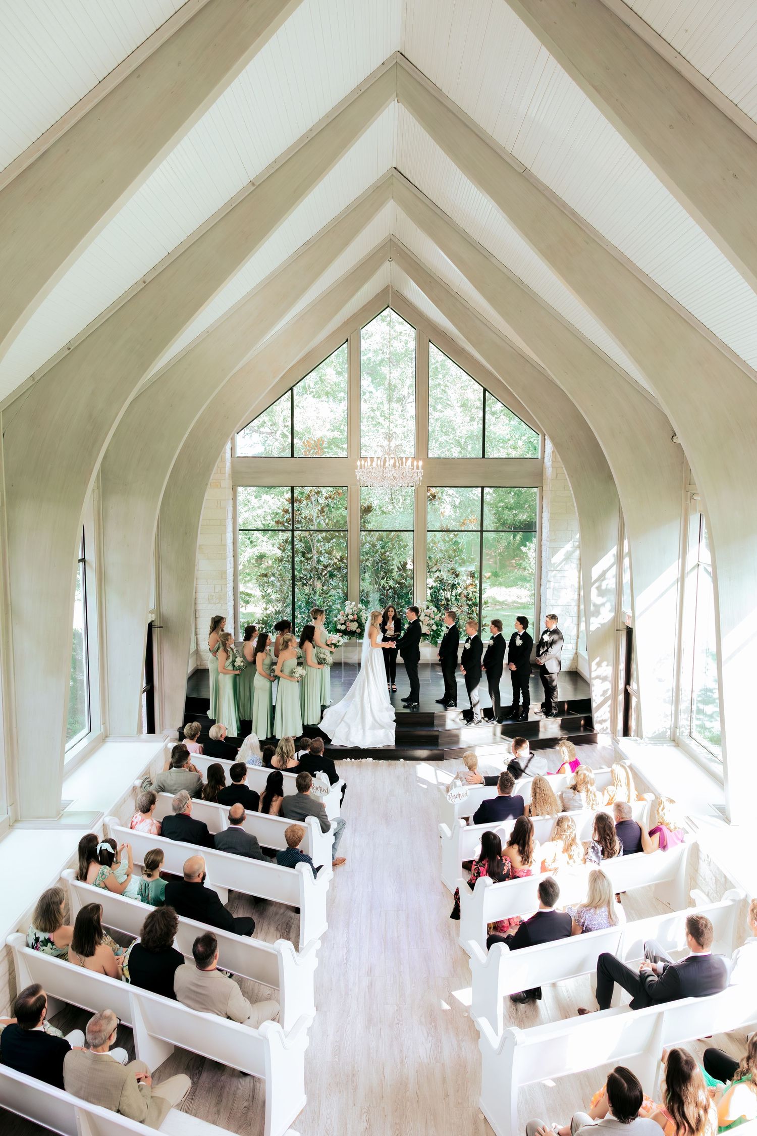 Wedding ceremony in a modern white chapel with vaulted ceilings and large windows overlooking greenery.