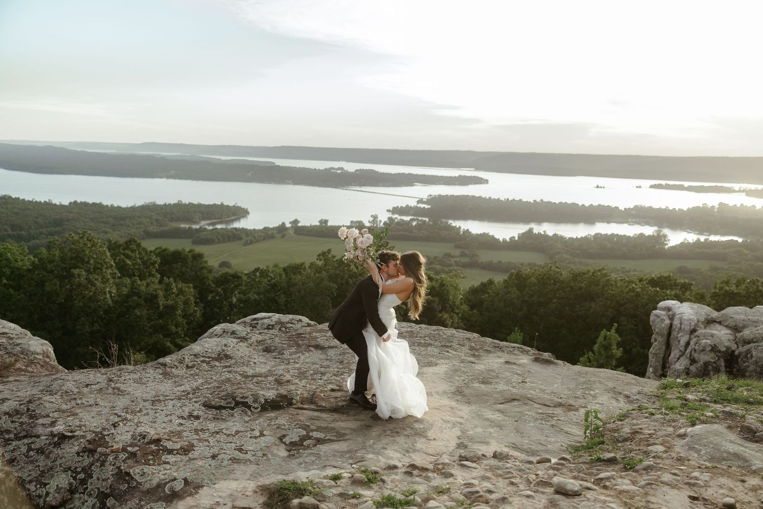 Bride and groom kissing on rocky hilltop overlooking scenic lake and lush green landscape at sunset.