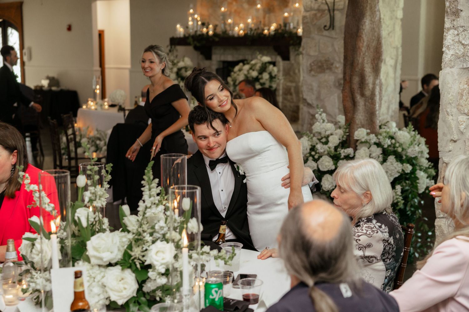 Bride in white strapless gown hugs guest at elegant wedding reception with white floral centerpieces and candlelight.