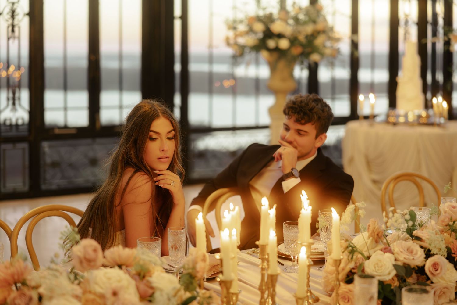 Couple seated at candlelit wedding reception table with floral arrangements and ocean view at dusk.