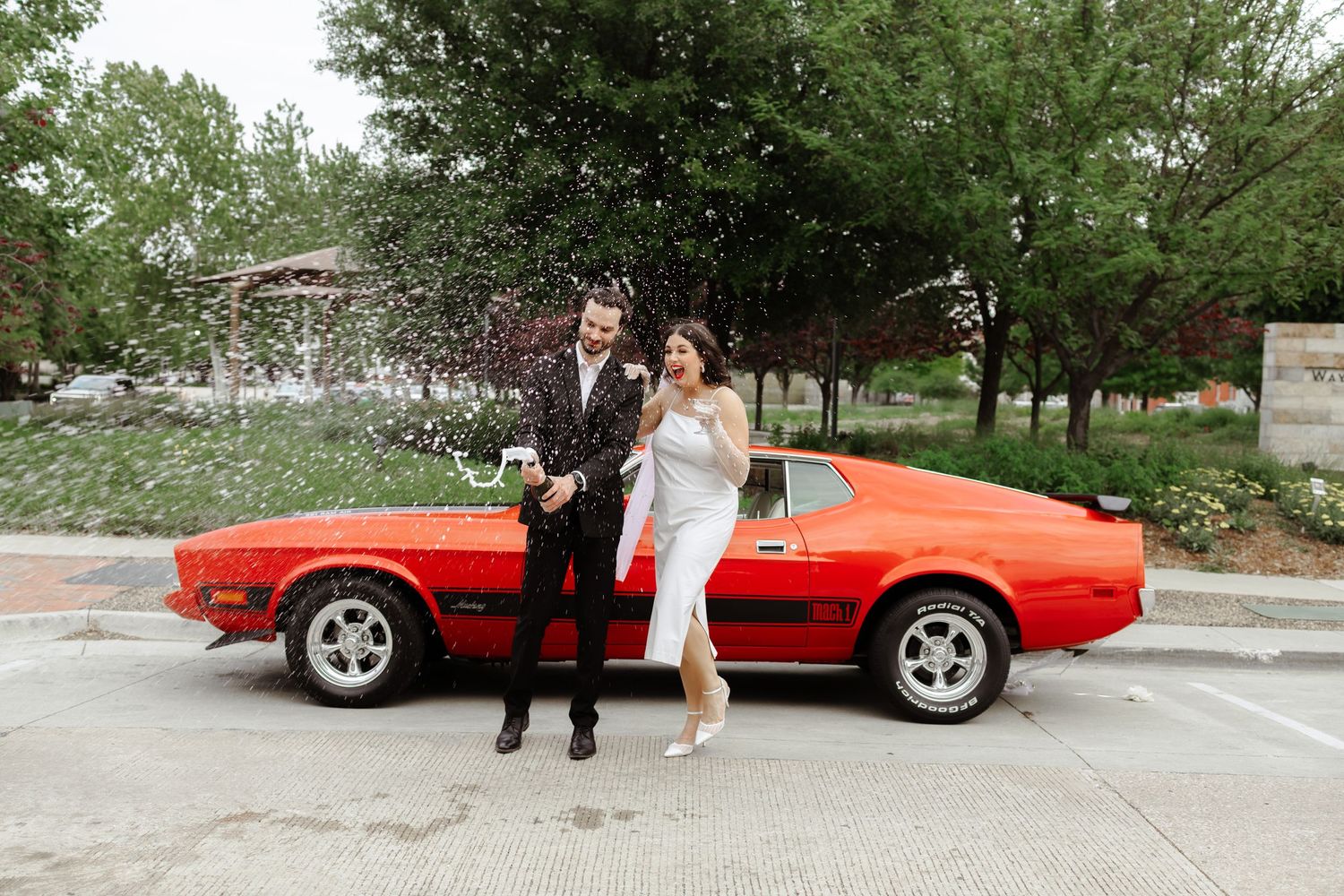 Bride and groom celebrating with champagne spray beside a classic red Ford Mustang in a parking lot.