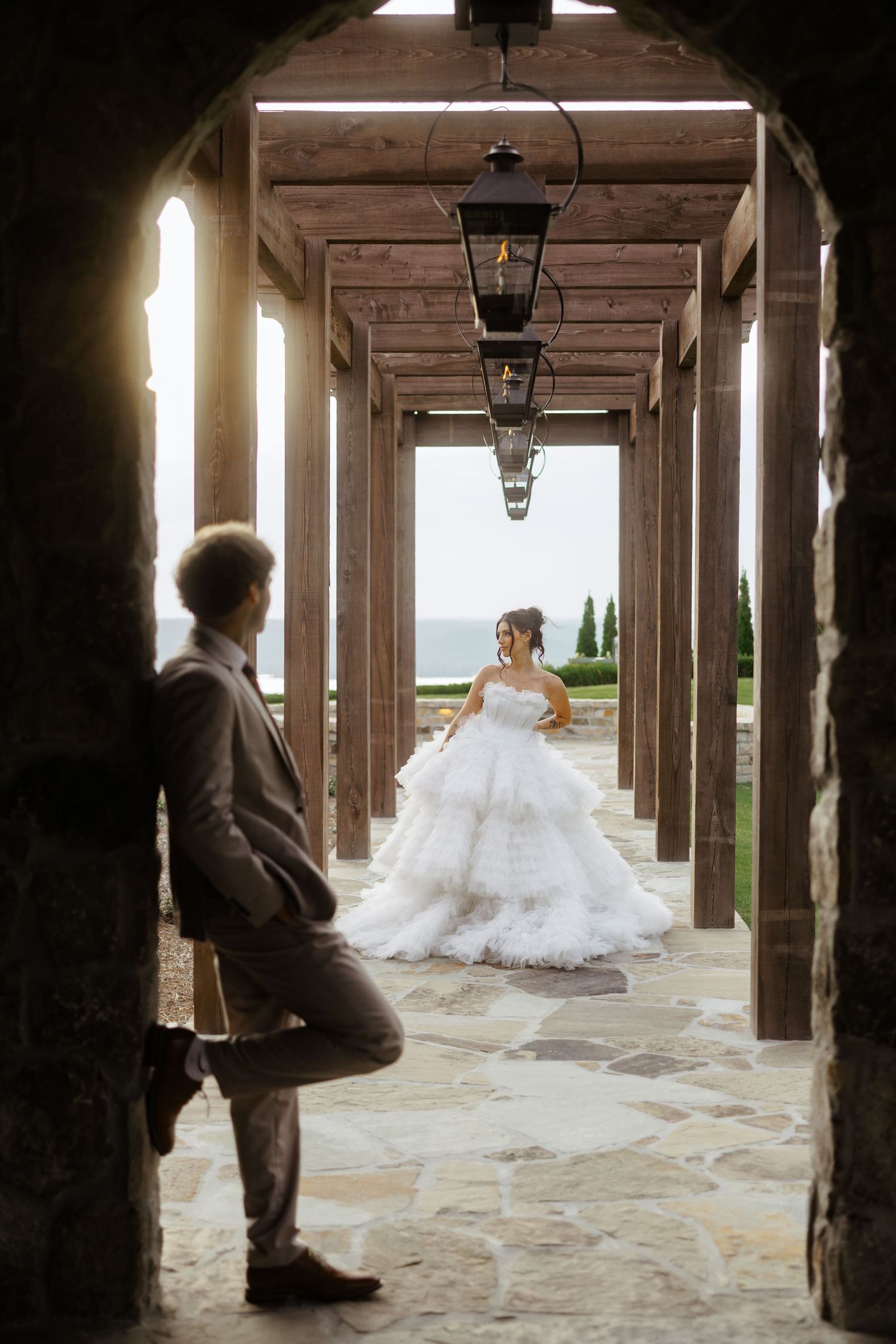 Bride in a white ballgown and groom in gray suit pose in a rustic stone colonnade with lanterns and garden views.