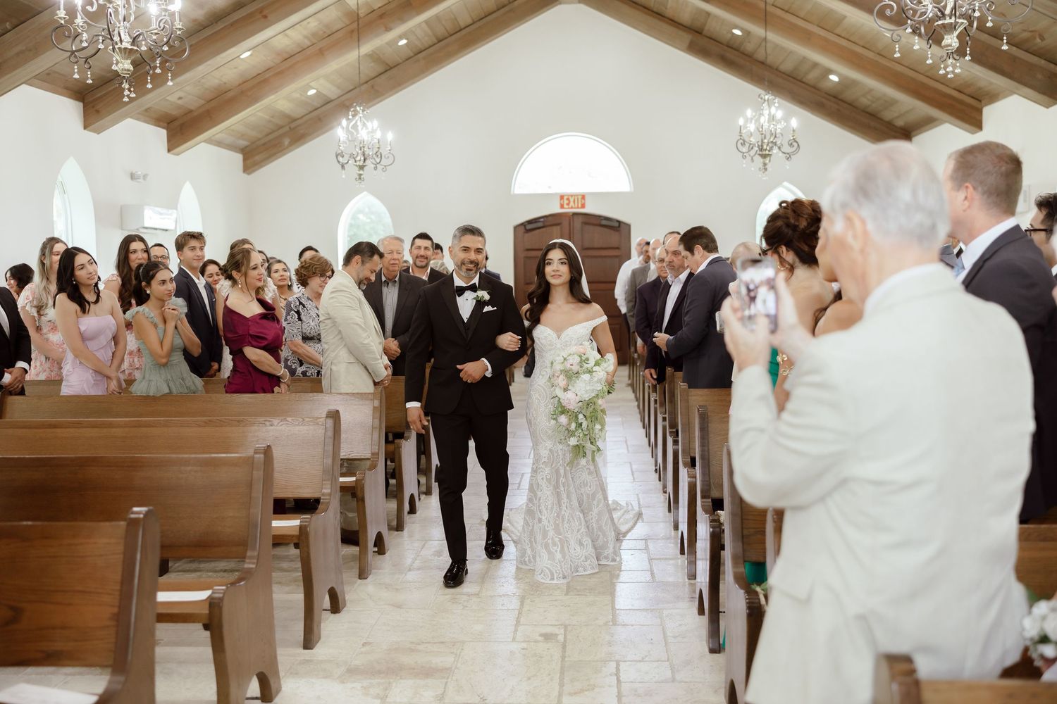 Bride walks down church aisle with escort during elegant wedding ceremony, guests standing in pews of white chapel.