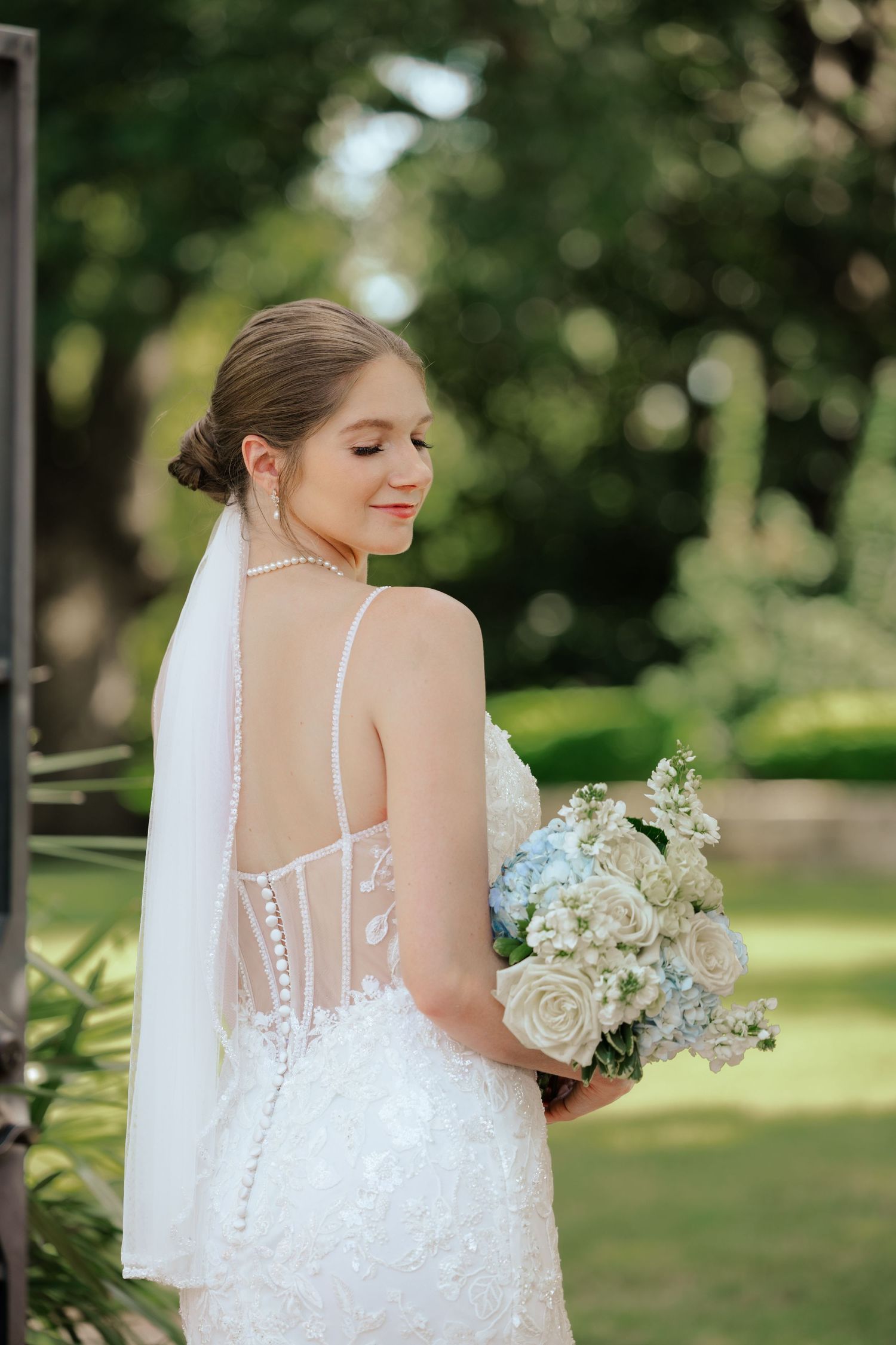 Bride in detailed lace gown holds bouquet of white roses in outdoor garden setting.