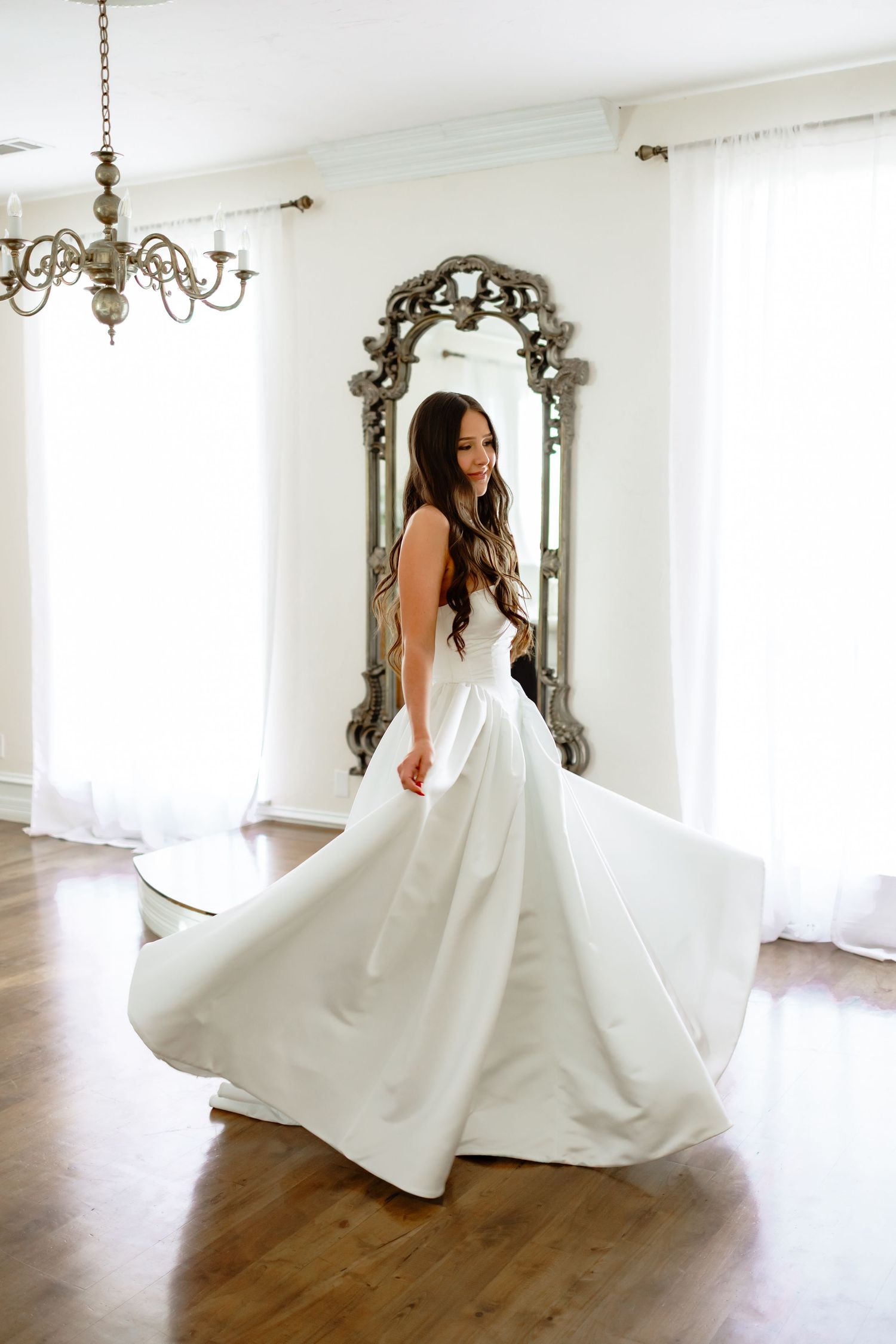 A bride in a flowing white wedding gown twirls in front of an ornate vintage mirror in a bright room with hardwood floors.
