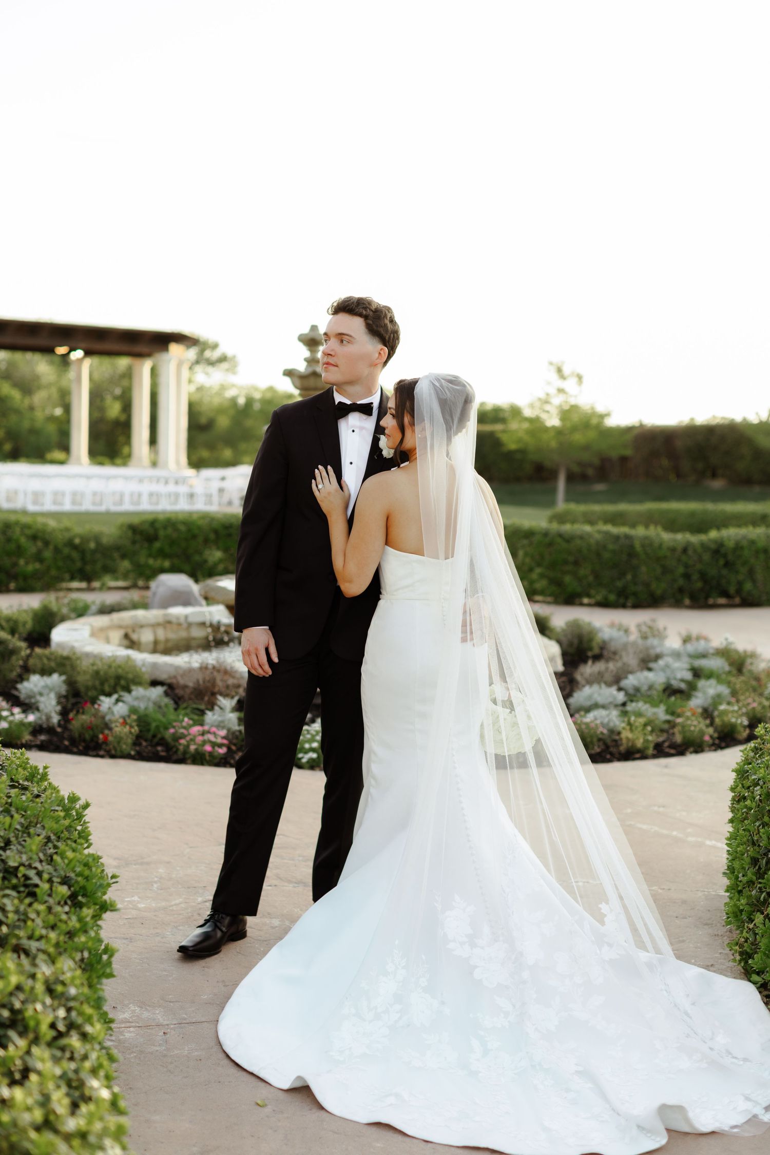 Bride in white mermaid gown with veil and groom in black tuxedo embrace in elegant garden wedding venue.