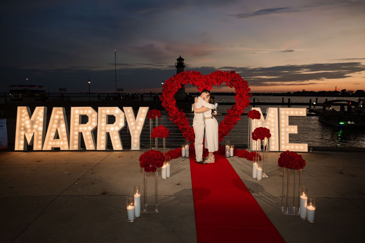 Romantic rooftop marriage proposal with illuminated MARRY ME letters, red heart backdrop, and couple at sunset.