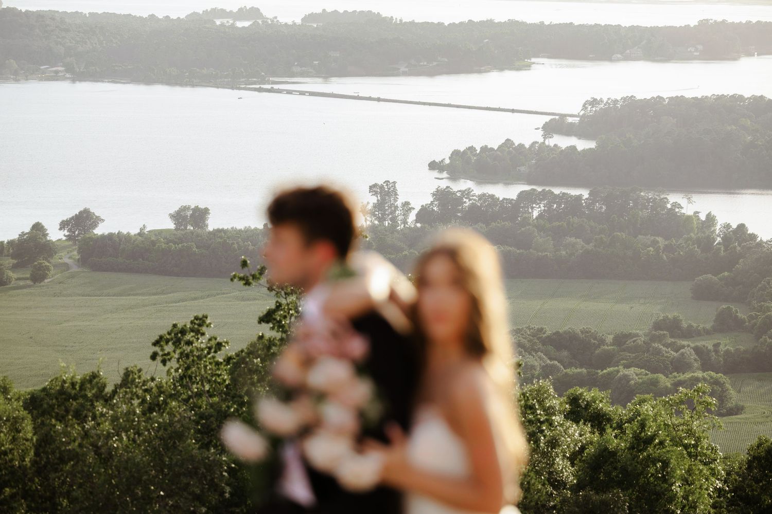 Blurred couple in wedding attire holding flowers with scenic lake and green landscape in soft background.