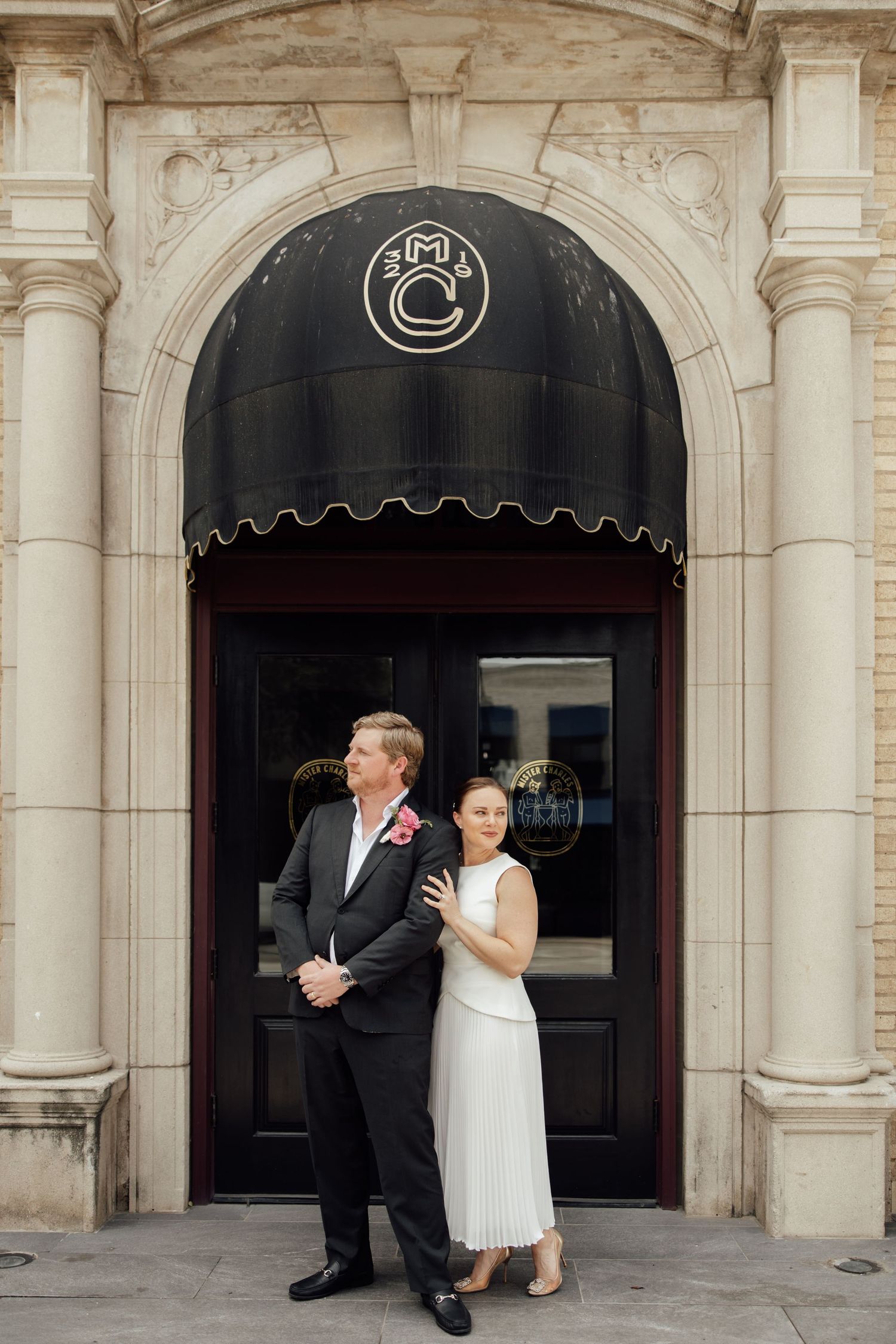 Couple in wedding attire posing in front of elegant arched doorway with black awning and ornate stone columns.