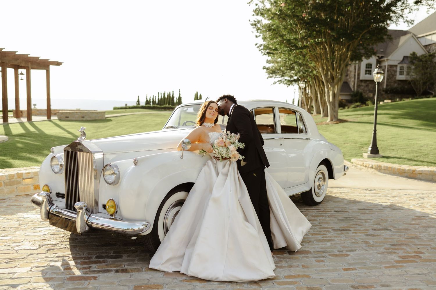 Bride and groom share a romantic kiss beside a classic white Rolls-Royce on a cobblestone driveway at sunset.