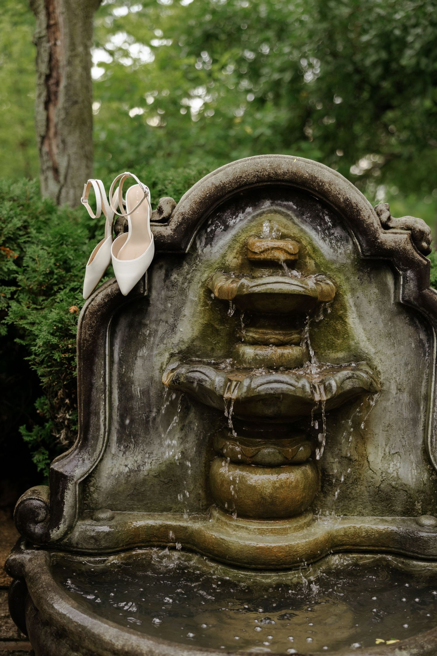 White bridal heels resting on an ornate stone garden fountain surrounded by lush green trees.