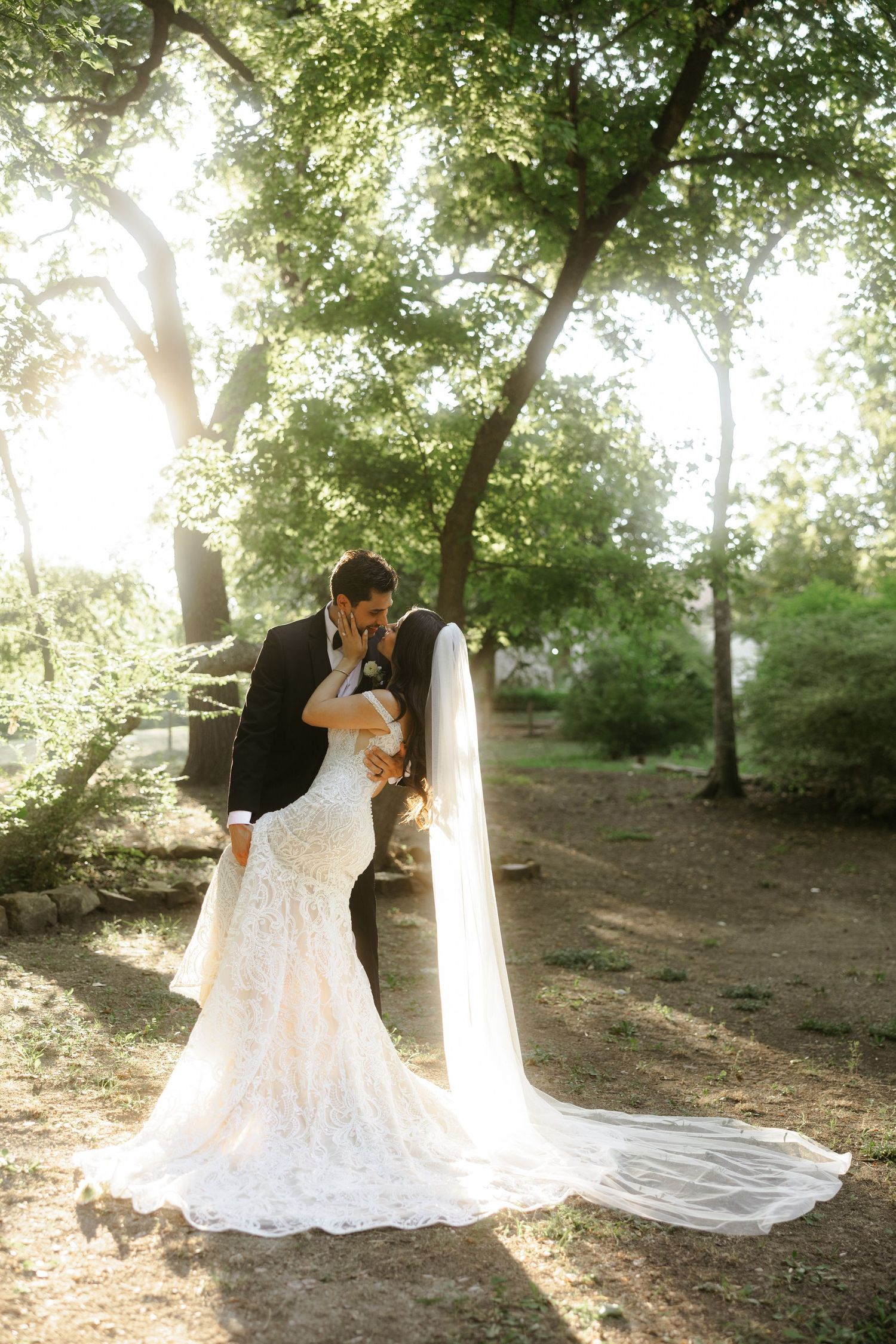Bride and groom share a romantic kiss in a sunlit forest, bride wearing a lace mermaid gown with cathedral veil.