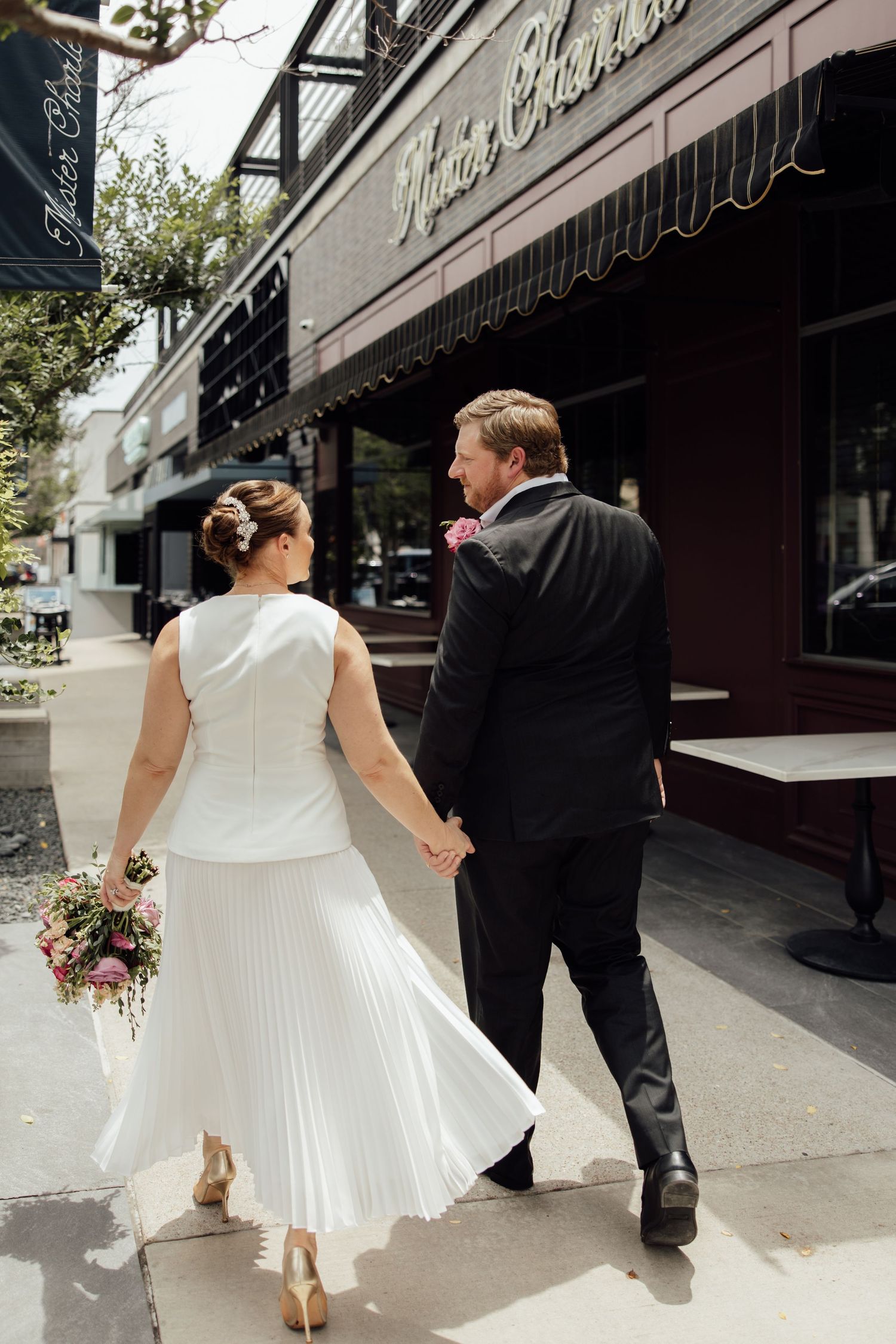 Bride and groom walk hand-in-hand along a city sidewalk, she in a white midi dress holding a bouquet, he in a black suit.