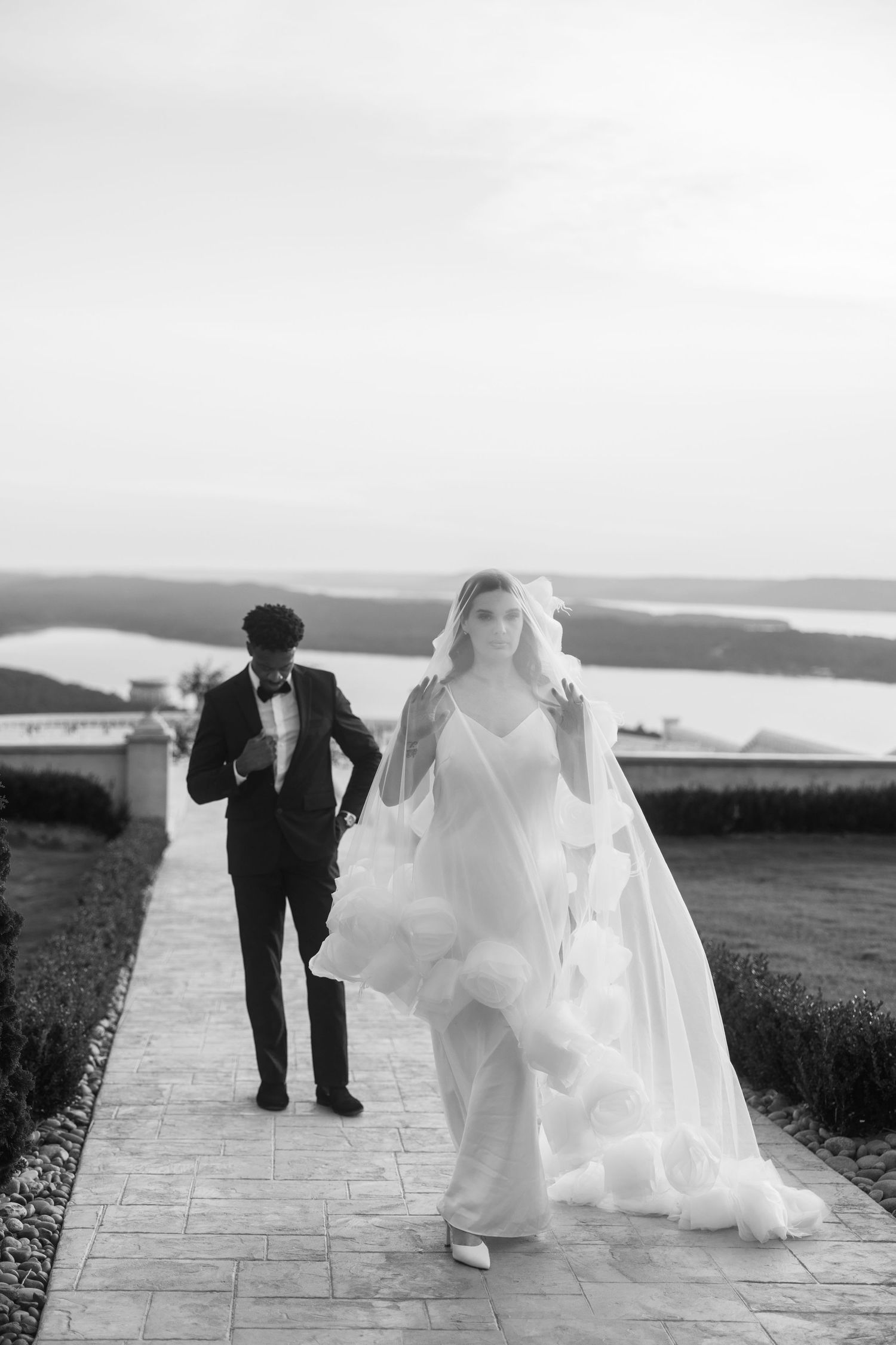 Black and white photo of bride in flowing gown and groom in tuxedo walking on stone path with scenic water view.