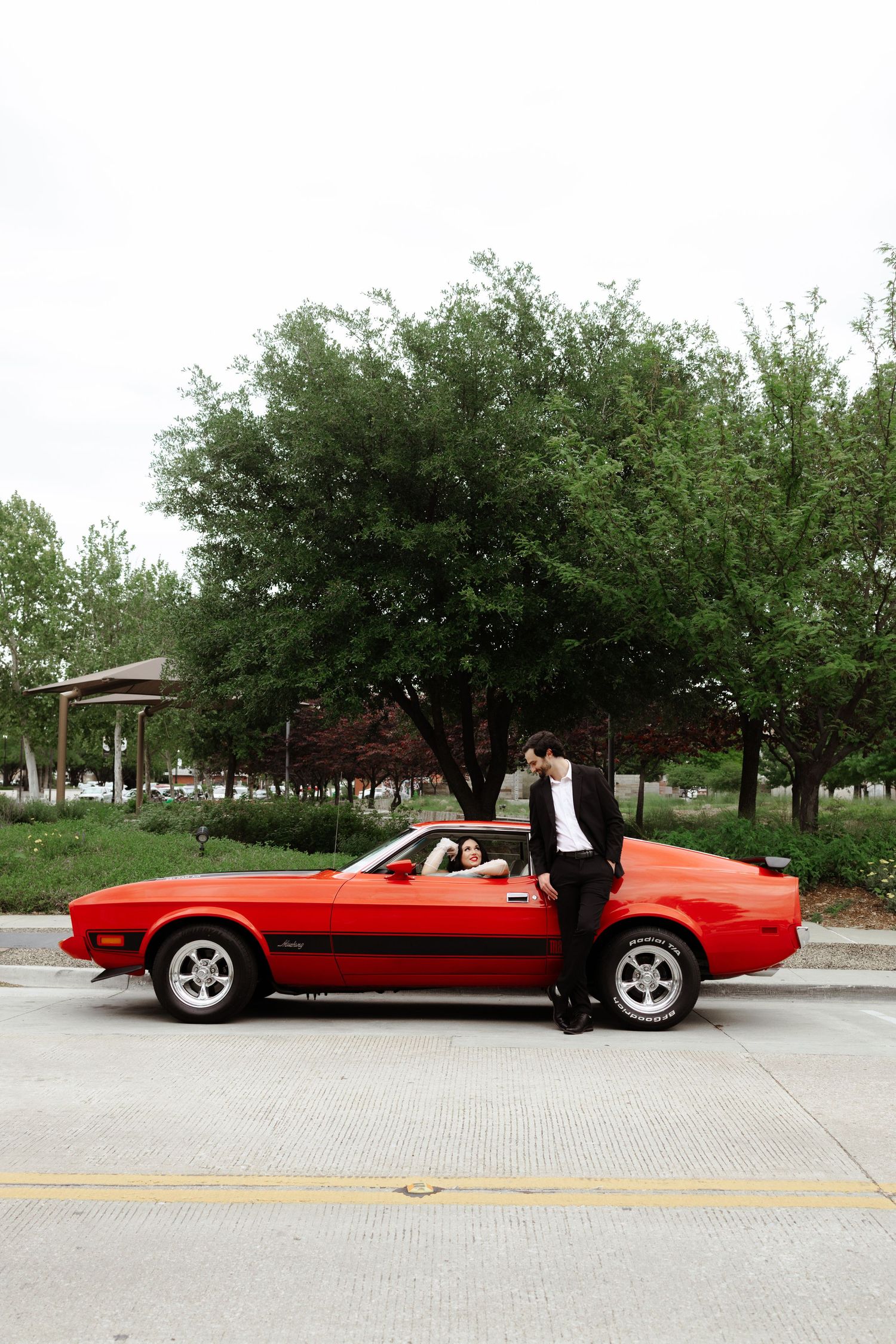 Couple posing with a classic red Ford Mustang on a street, man in suit leaning on car door.