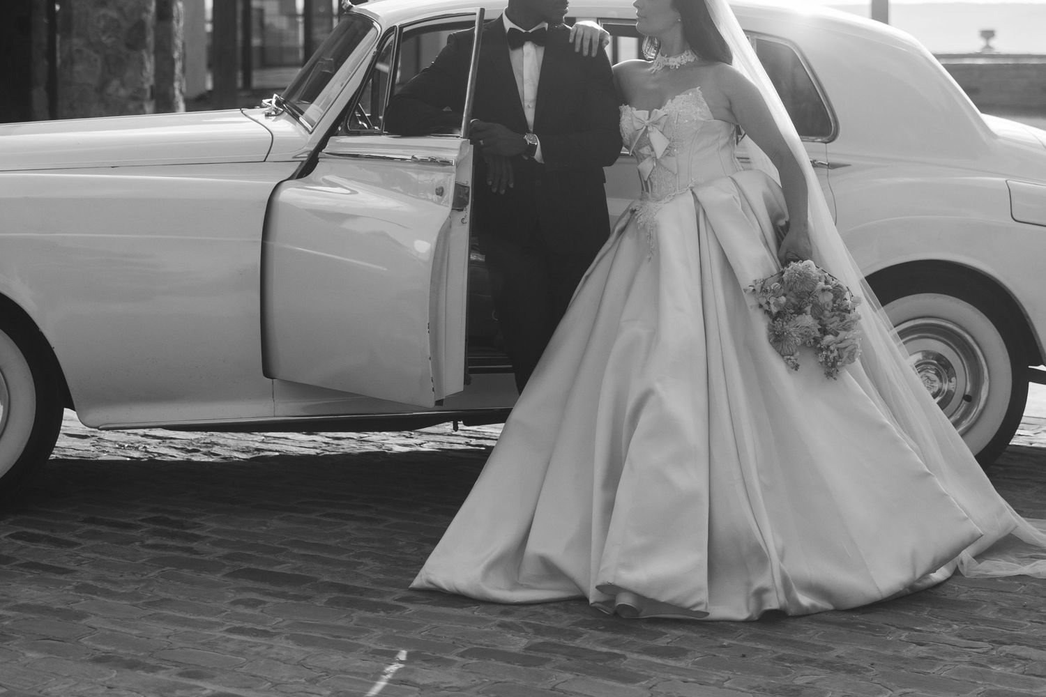 Black and white photo of a bride in a ball gown exiting a vintage white Rolls-Royce with a groom in a tuxedo.