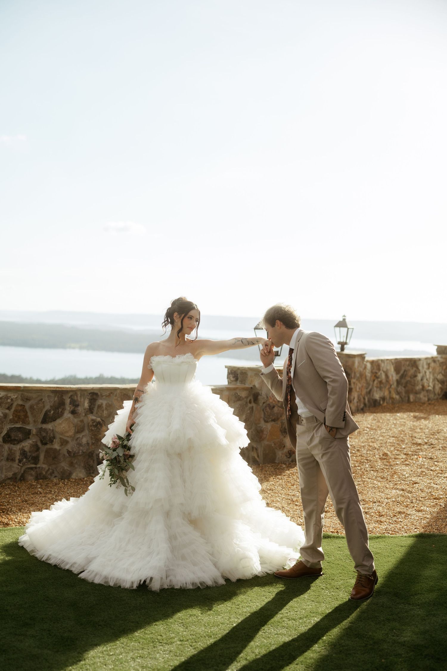 Groom in gray suit kisses bride's hand outdoors, bride wearing ruffled white ballgown holding bouquet with ocean view.