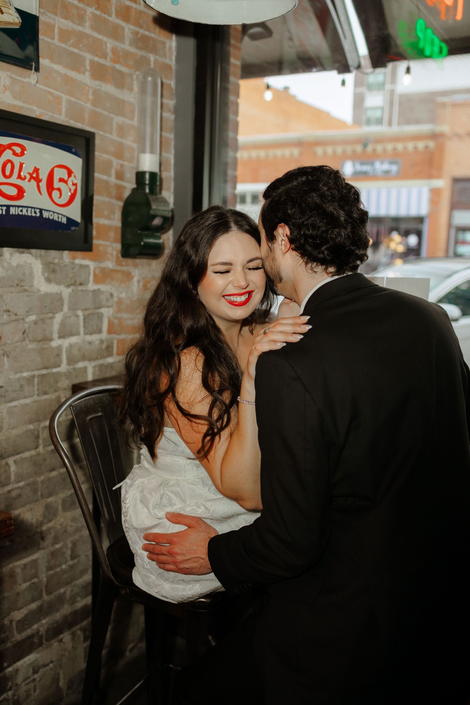 Groom in black suit kissing bride on cheek at a bar, bride laughing with red lips and white wedding dress.