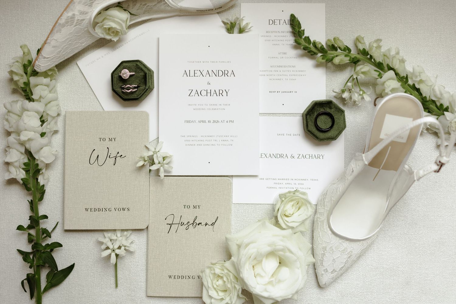 Elegant wedding stationery flat lay with ivory invitations, vow books, floral greenery, ring box, and white heels.