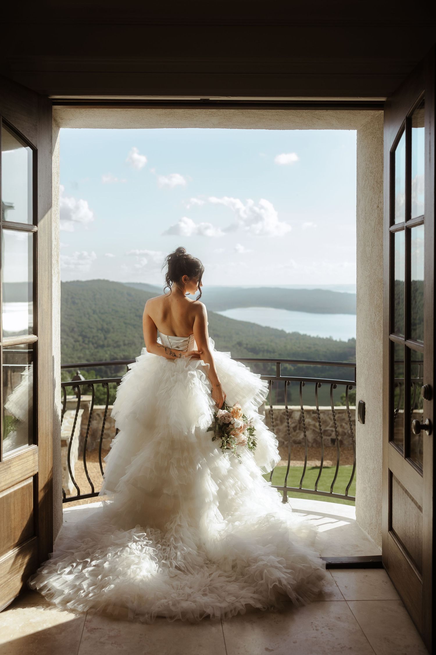 Bride in a white ruffled gown stands in open doors overlooking a scenic lake and forest landscape.