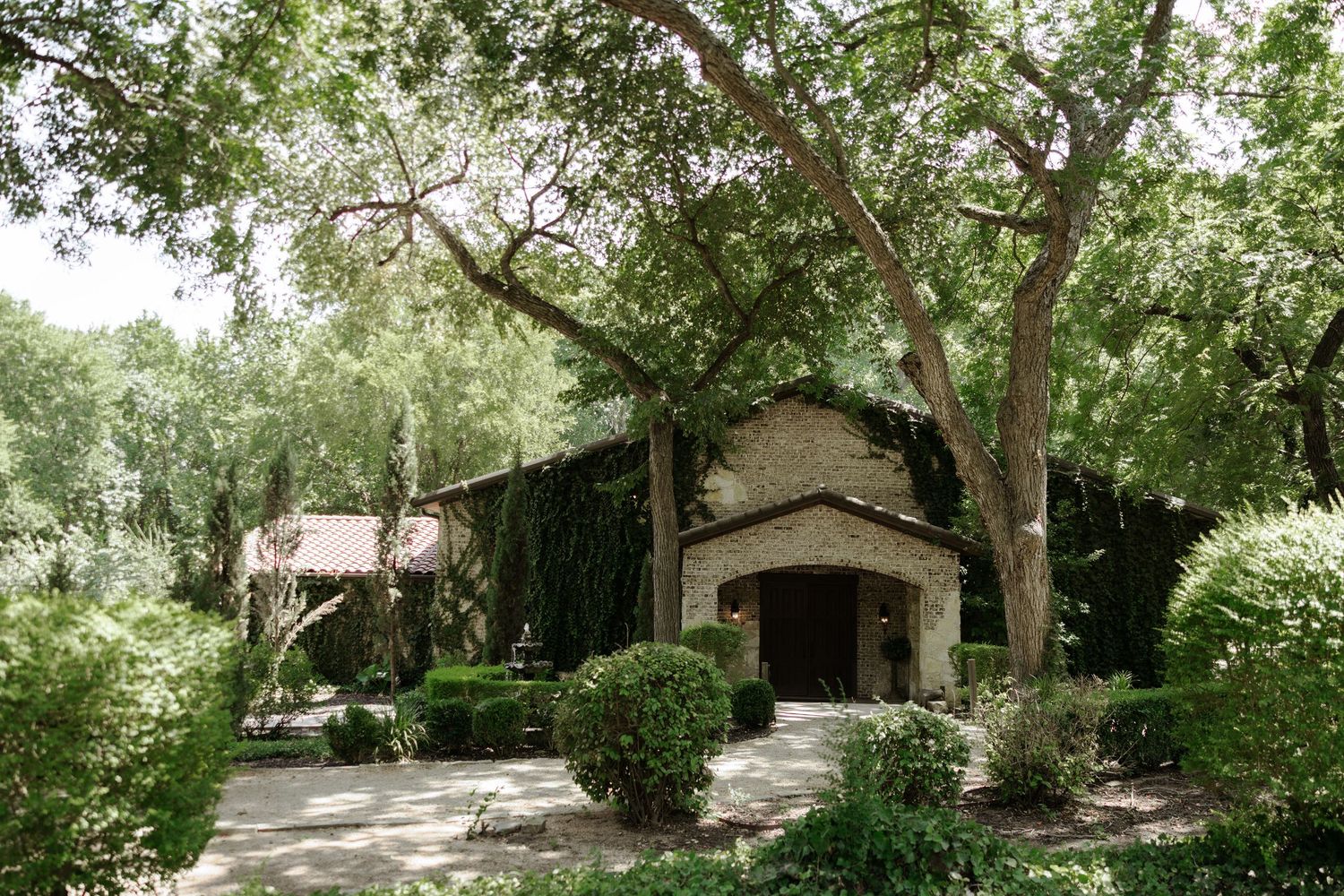 Stone building surrounded by lush green trees and manicured shrubs along a shaded driveway.