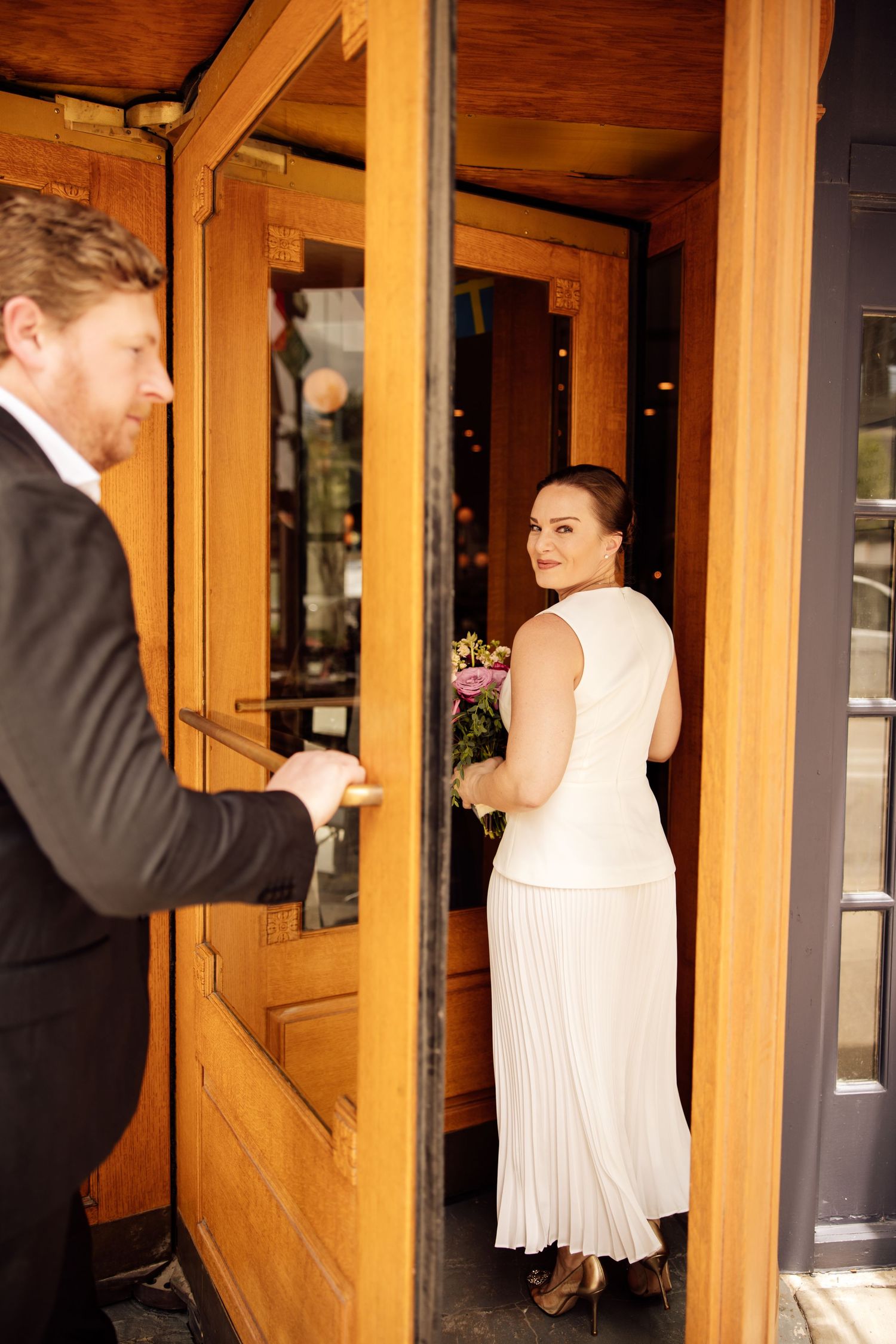 Groom in black suit holds door open for smiling bride in white dress holding bouquet at restaurant entrance.