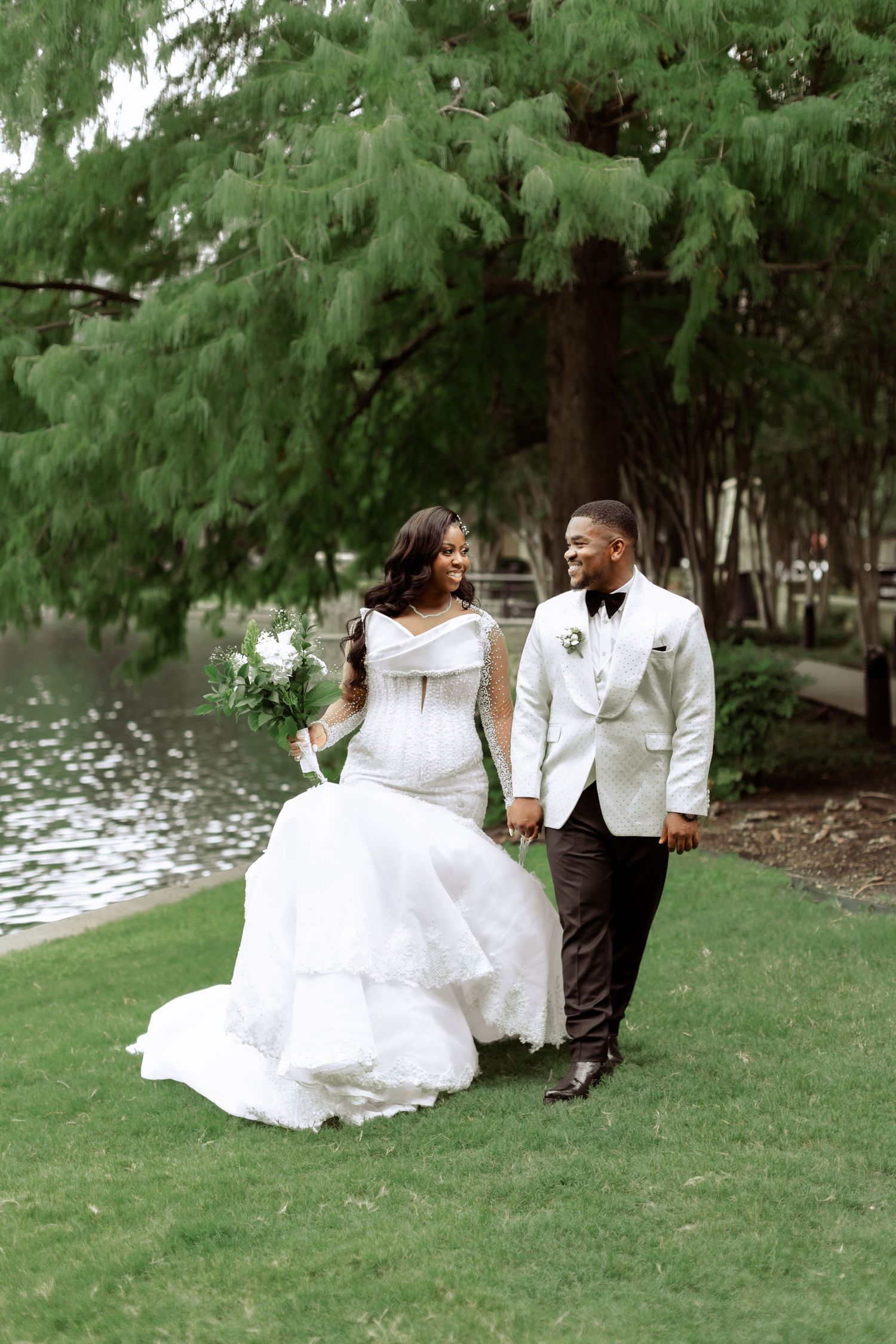 A romantic wedding couple sits together on grass near a serene lake surrounded by evergreen trees.