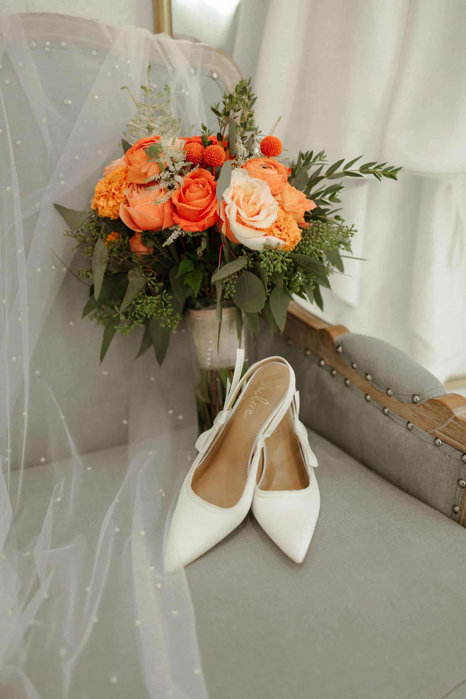 Wedding accessories featuring white pointed-toe heels and coral orange rose bouquets arranged on a light gray surface.